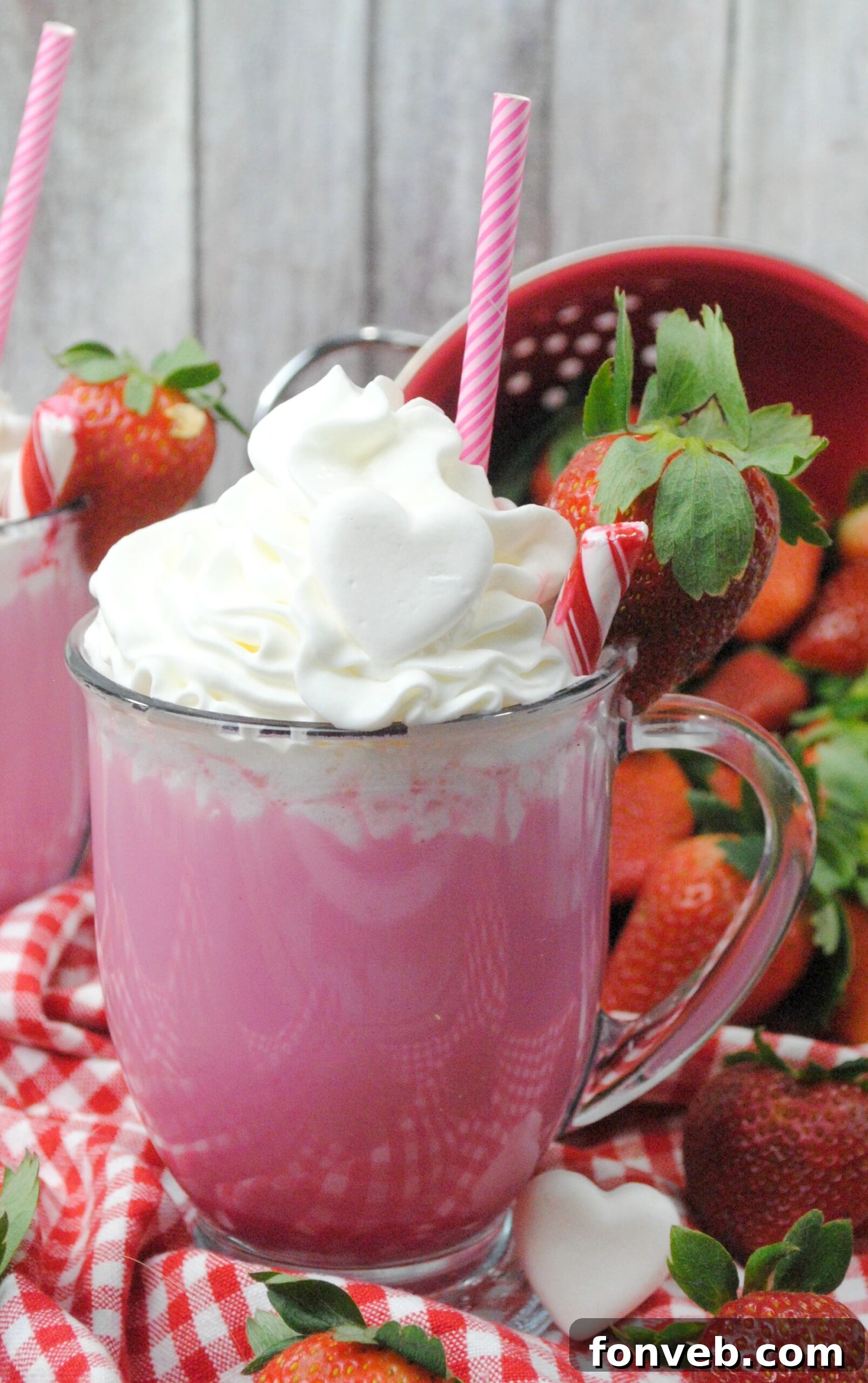 Two mugs of Pink Velvet Hot Chocolate, one topped with whipped cream and sprinkles, next to fresh strawberries on a rustic wooden surface.
