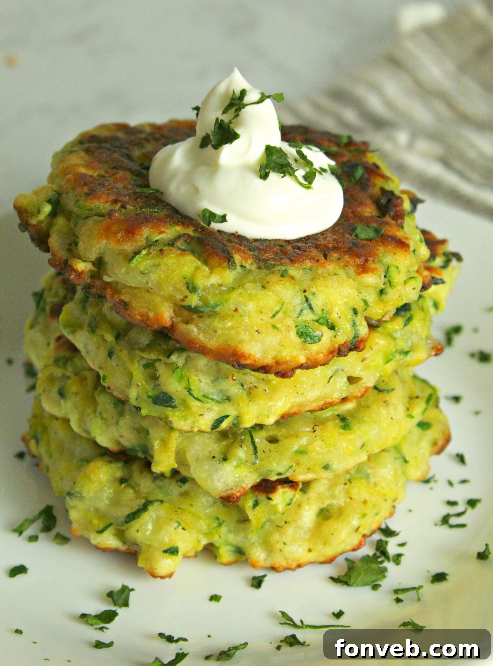 Two cheesy zucchini pancakes on a white plate with sour cream and fresh herbs