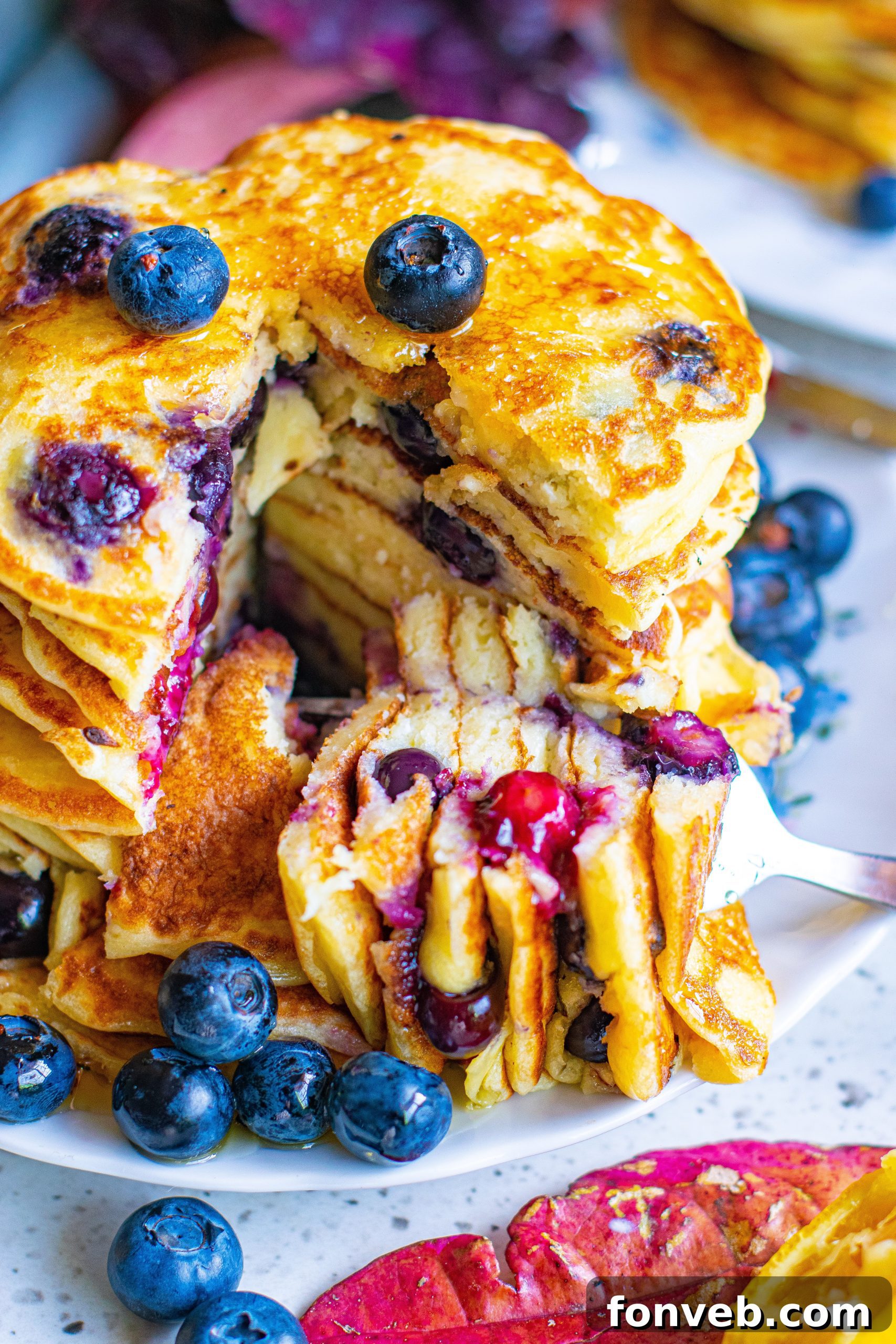 A tall stack of golden blueberry cream cheese pancakes, generously drizzled with maple syrup and topped with fresh blueberries.