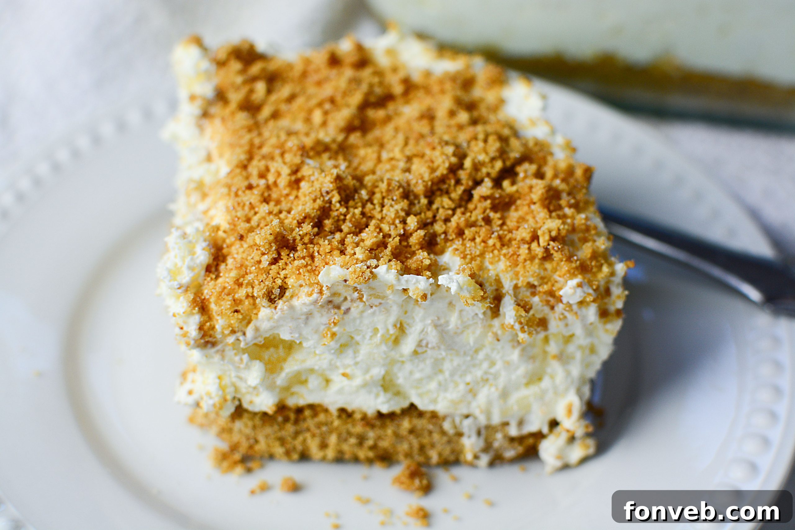 A freshly cut square of pineapple cream dessert, showing distinct layers of crust, cream cheese filling, and pineapple whipped topping, against a rustic background.