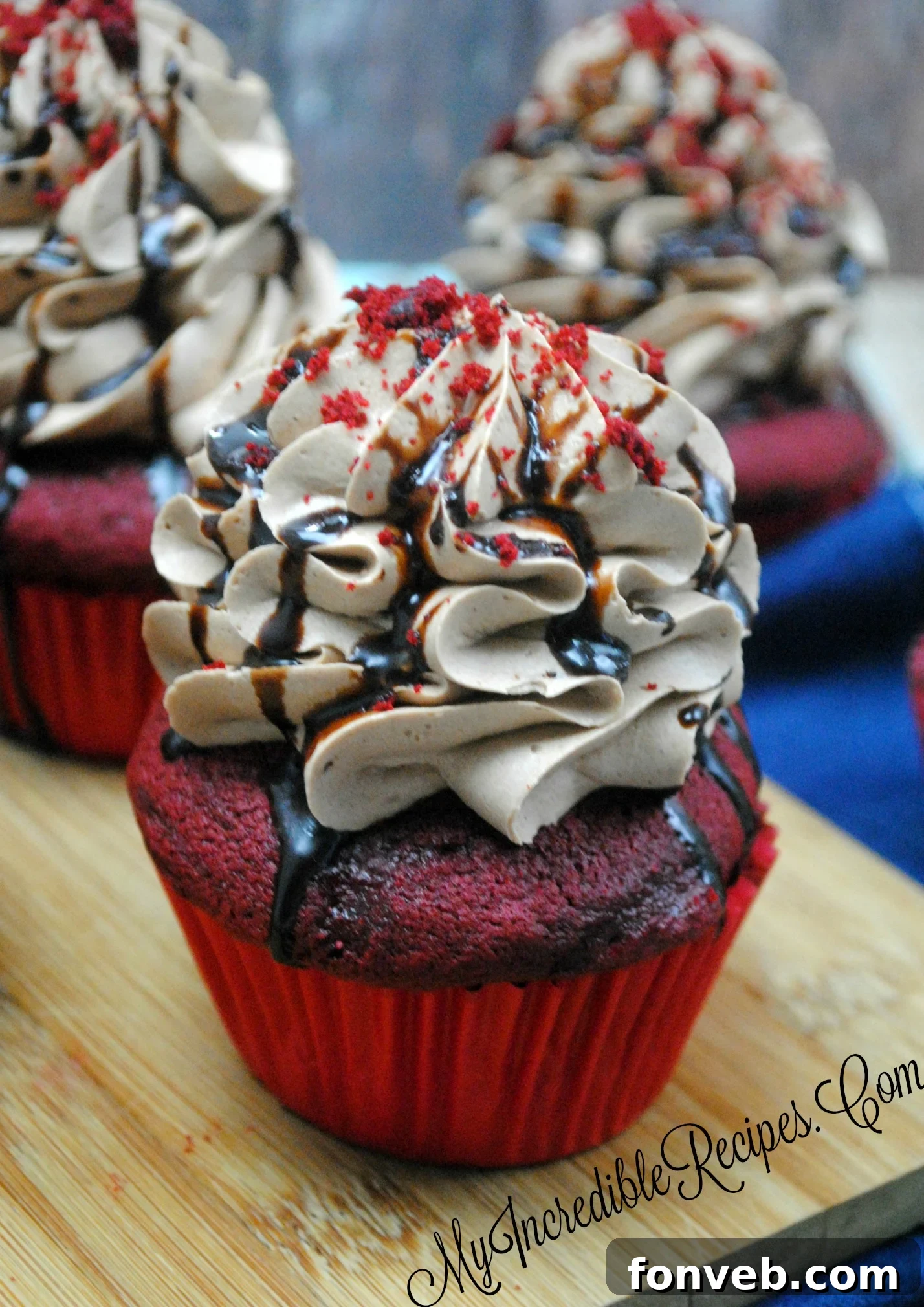 A frosted Red Velvet cupcake being drizzled with hot fudge sauce
