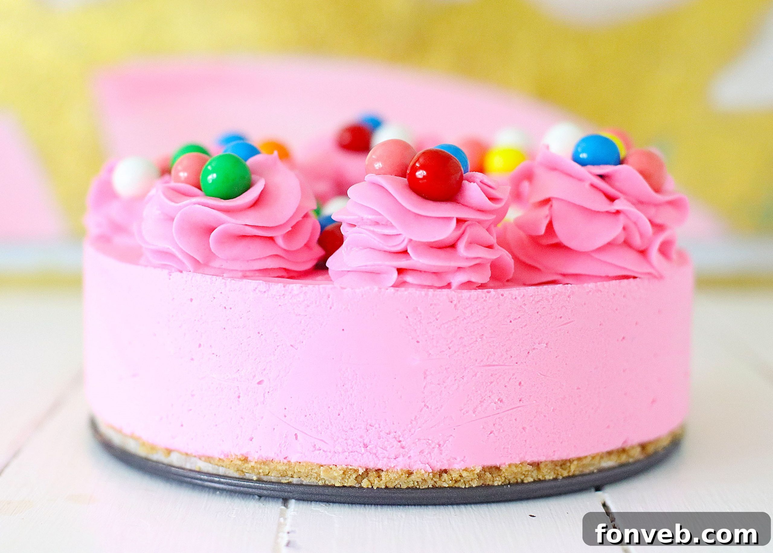 A hand gently folding colorful gumballs into a bowl of creamy, pink cheesecake batter, preparing it for the crust.
