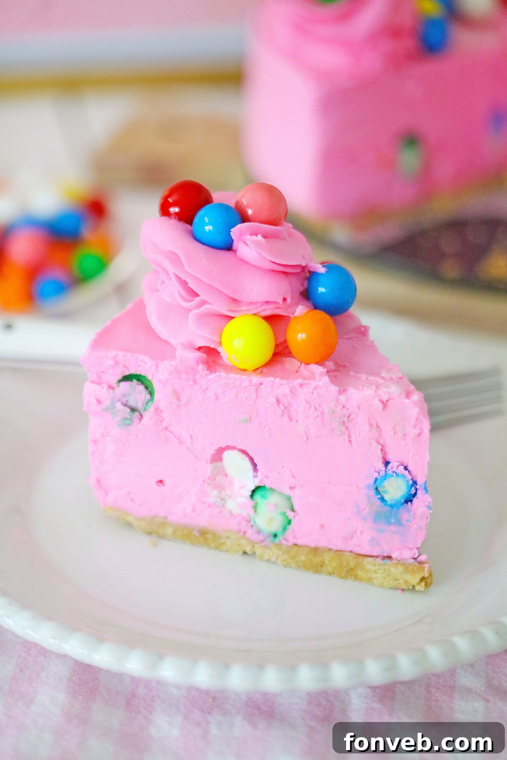 A close-up of a generous slice of bubblegum cheesecake on a white plate, revealing the gumballs and the thick, creamy pink filling.