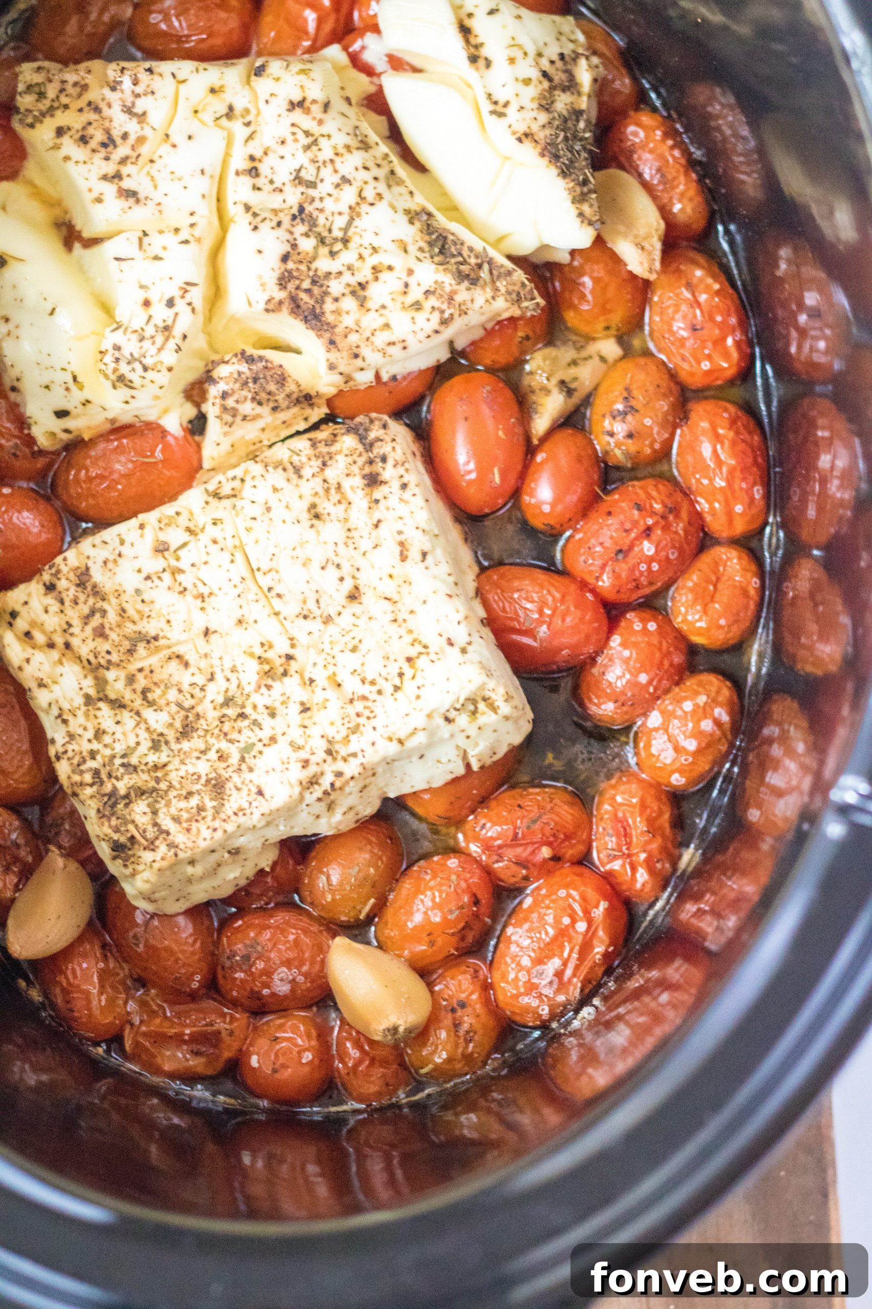 A close-up view of the slow cooker filled with melting feta and cream cheese over cherry tomatoes and garlic, before stirring.