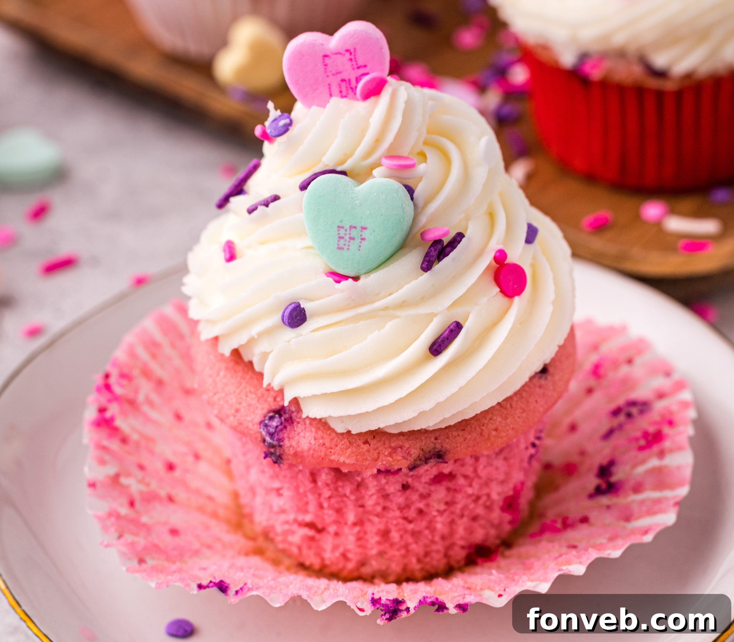 Blushing Velvet Vanilla Cupcakes 10 Close-up of a single pink velvet cupcake adorned with white vanilla buttercream, pink sprinkles, and a small heart candy.