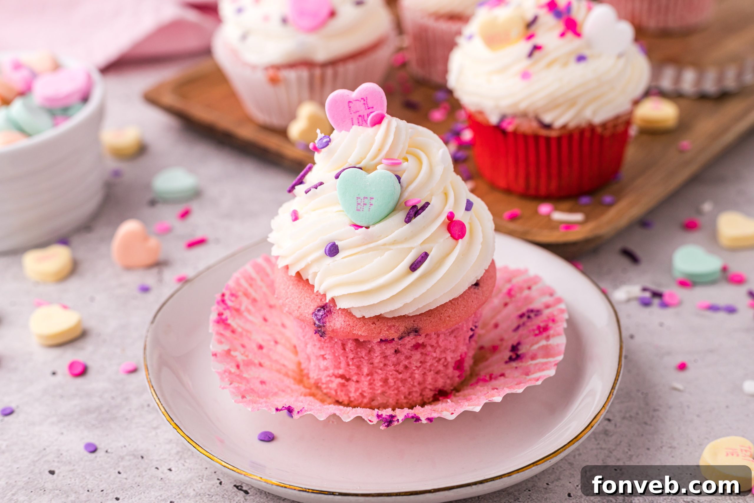 Blushing Velvet Vanilla Cupcakes 12 Artistic shot of a single pink velvet cupcake with swirled vanilla buttercream, adorned with a heart candy and sprinkles, on a rustic surface.