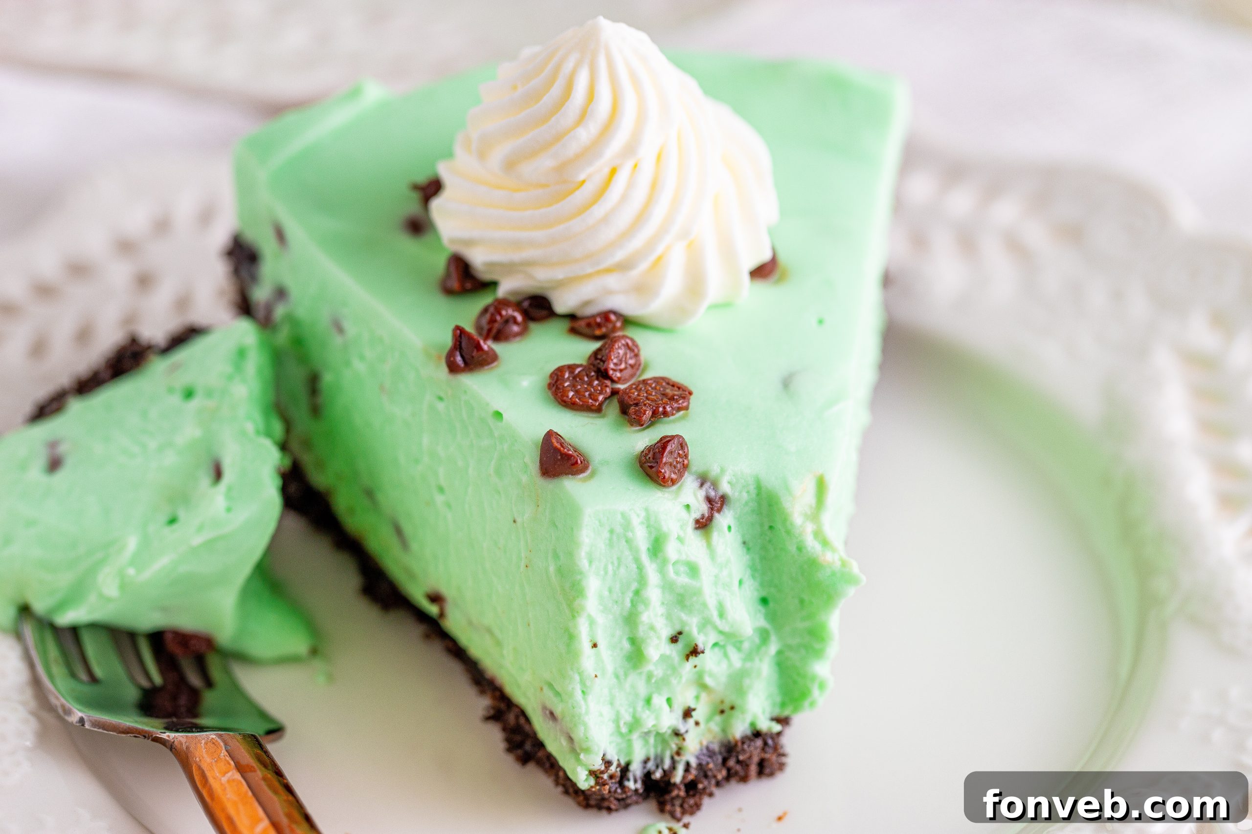 A close-up of a beautifully cut slice of No Bake Mint Chocolate Chip Cheesecake on a white plate, with a spoon taking a bite.