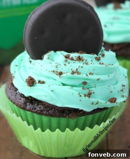 A freshly frosted Thin Mint cupcake, topped with a whole Thin Mint cookie and crushed cookie pieces, against a blurred background.