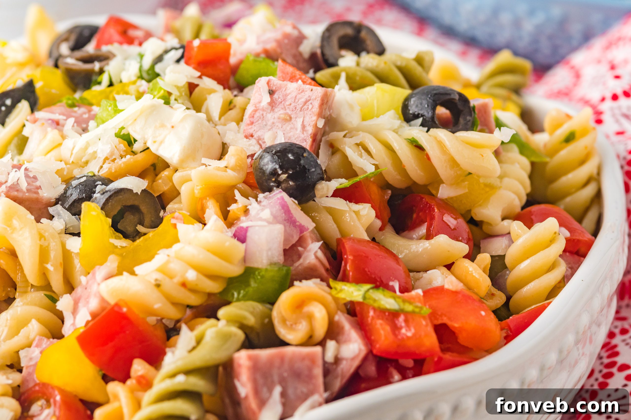 A wide shot of the Italian Pasta Salad in a large white bowl, surrounded by fresh ingredients like bell peppers and herbs, indicating its freshness and appeal.