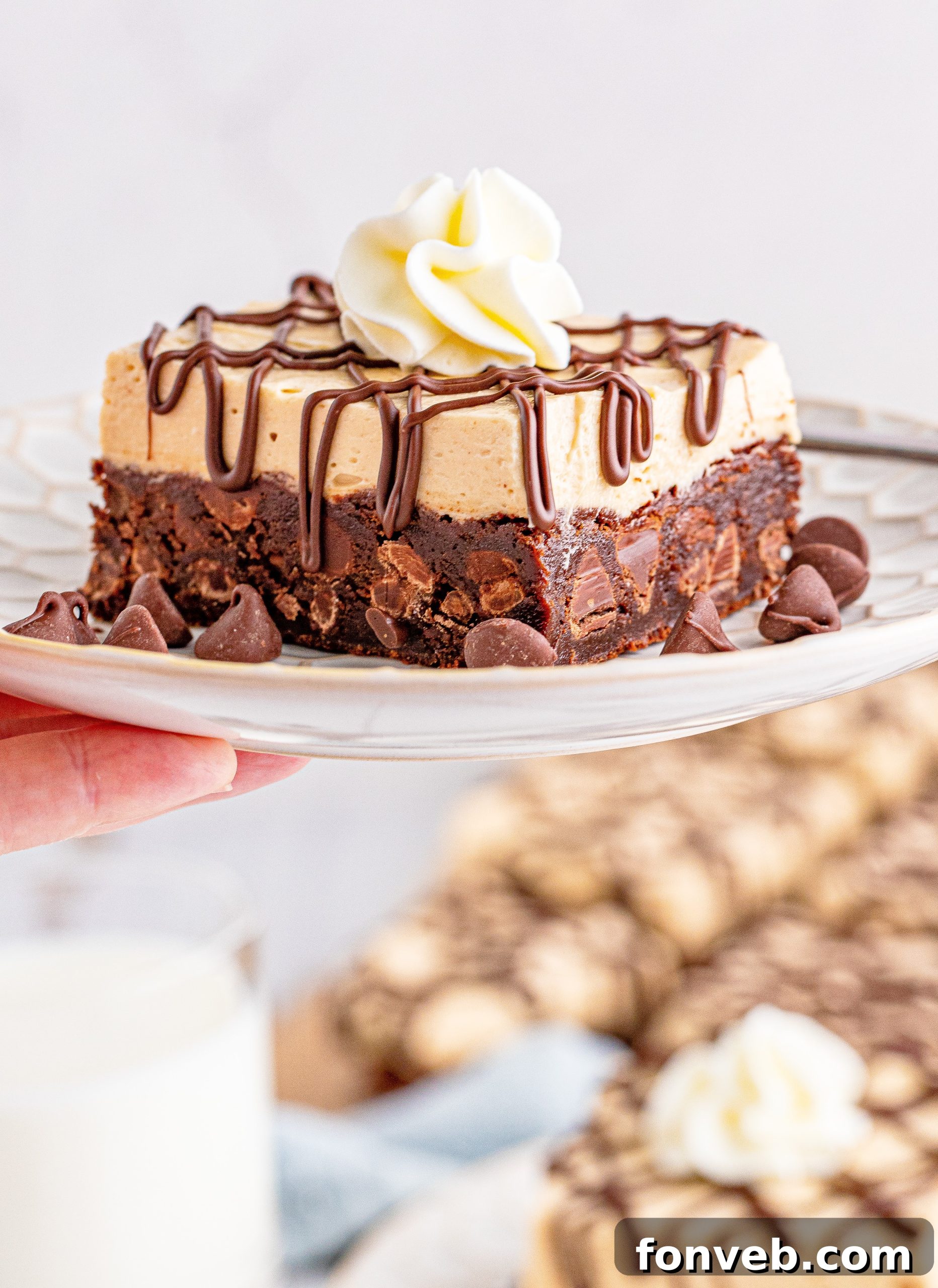 a hand holding up a plate with Peanut Butter Brownies on a white place drizzeled with chocolate and topped with whipped cream
