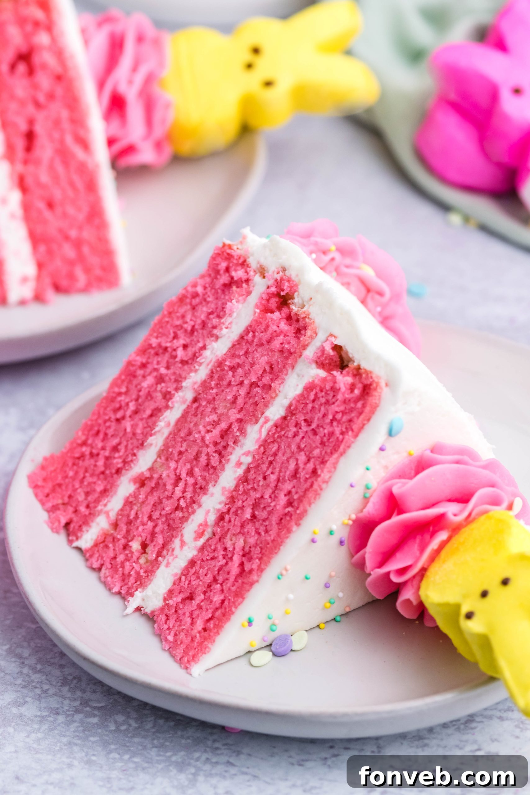 Pink Vanilla layered cake with white frosting and topped with yellow peeps for easter on a white plate with two other slices next to it