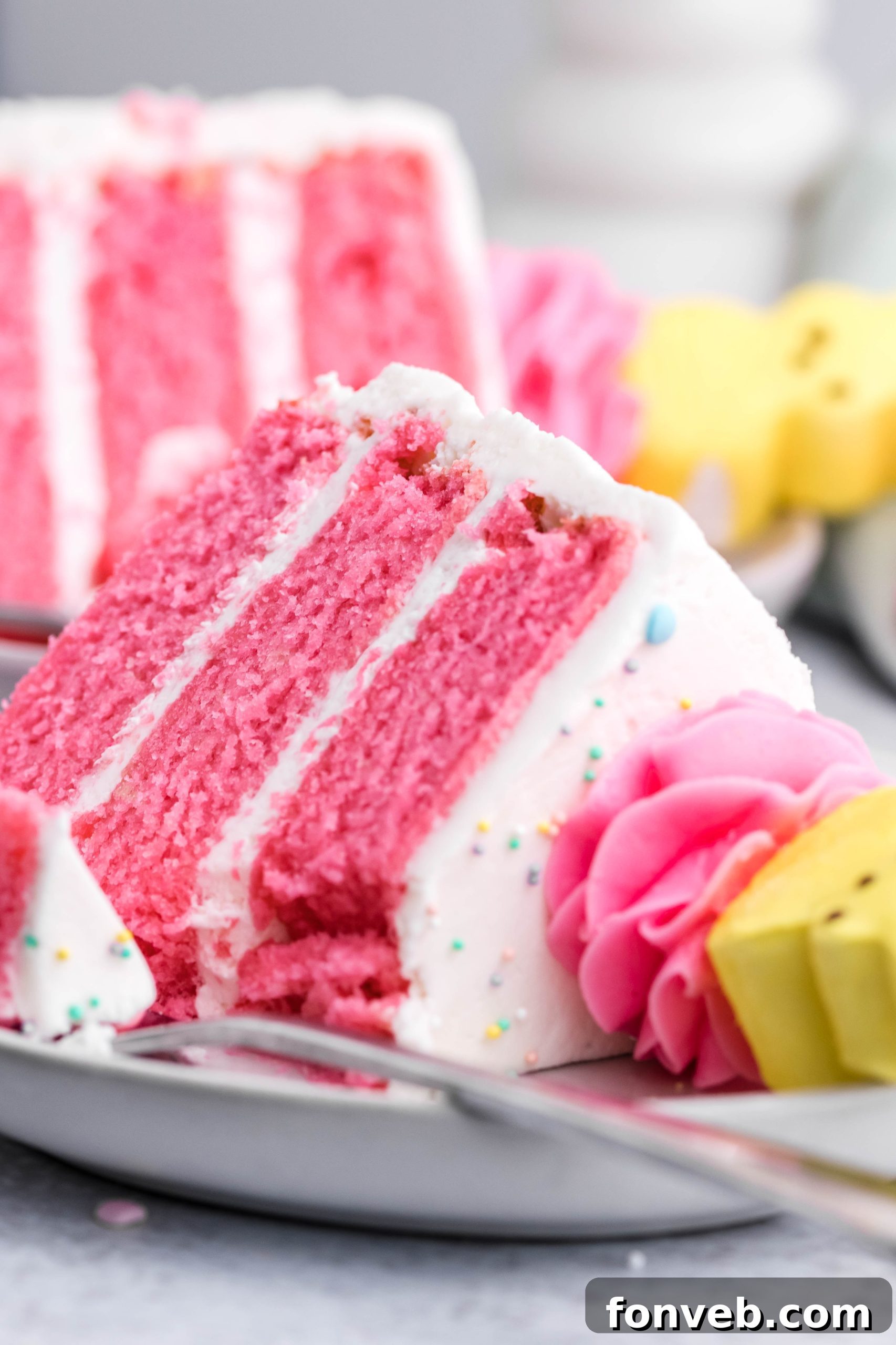 Pink Vanilla layered cake with white frosting and topped with yellow peeps for easter on a white plate with a bit gone