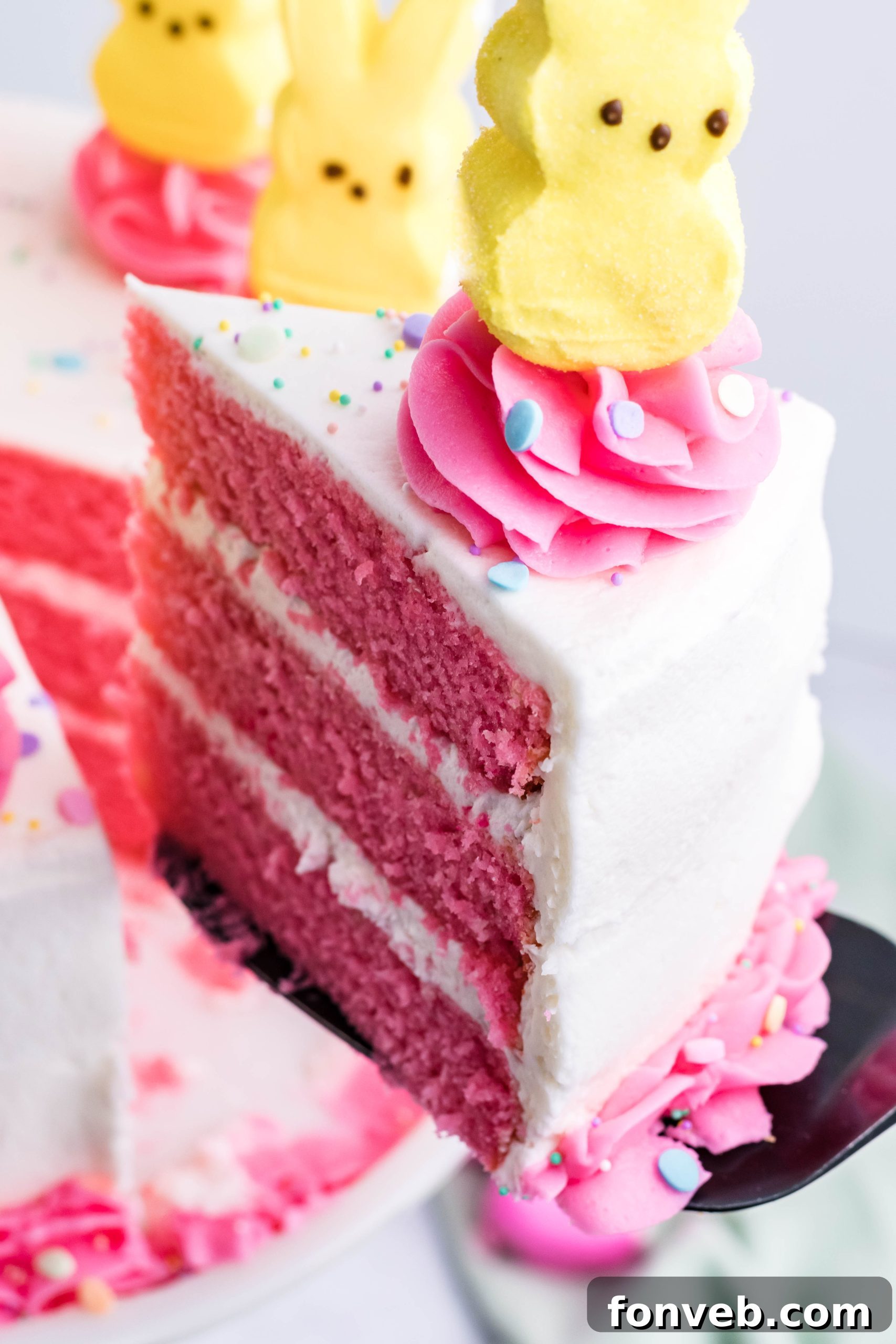 Pink Vanilla layered cake with white frosting and topped with yellow peeps for easter on a white cake stand being lifted up by a black cake cutter
