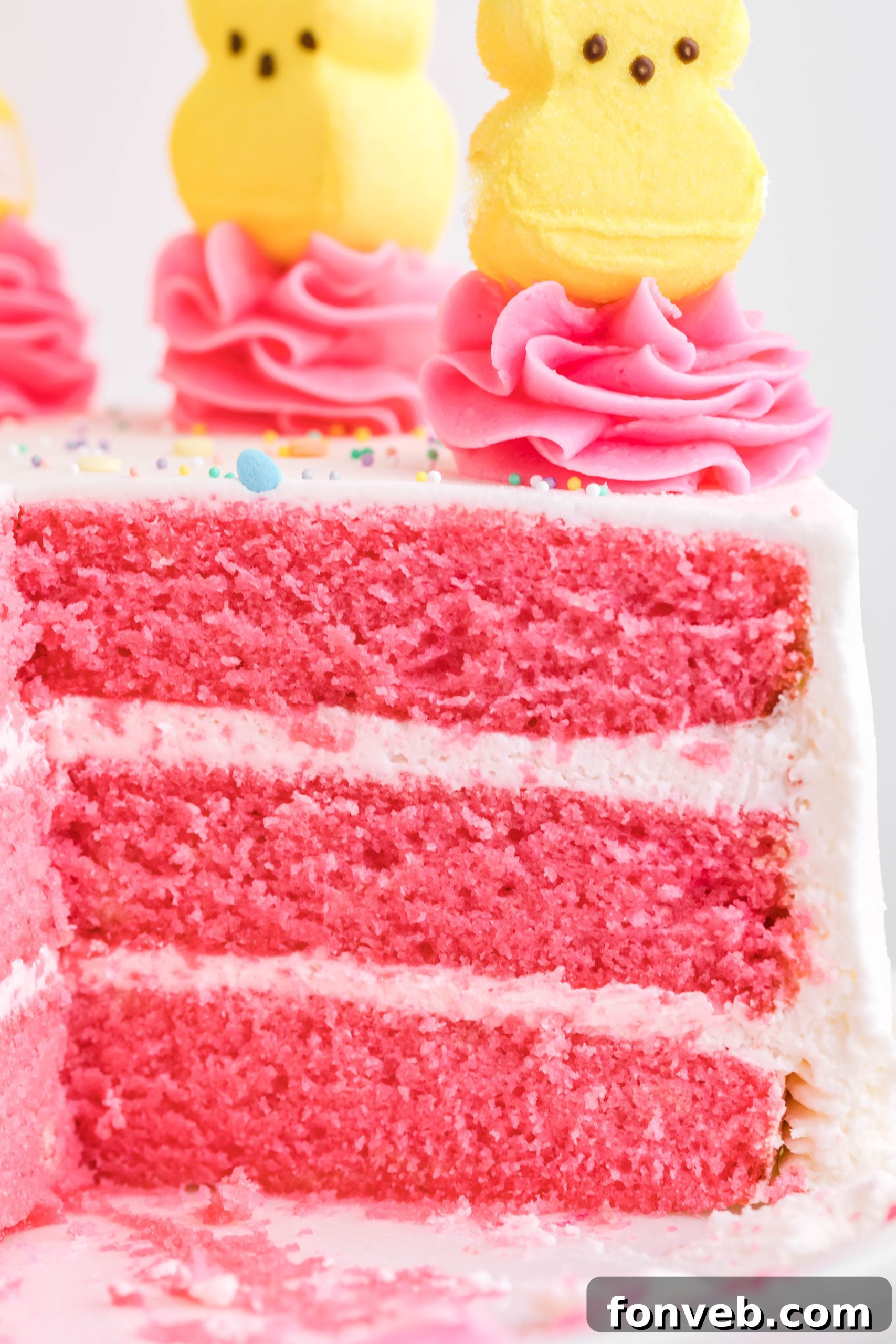 Pink Vanilla layered cake with white frosting and topped with yellow peeps for easter on a white cake stand from the side to show a close up of all the hot pink layers 