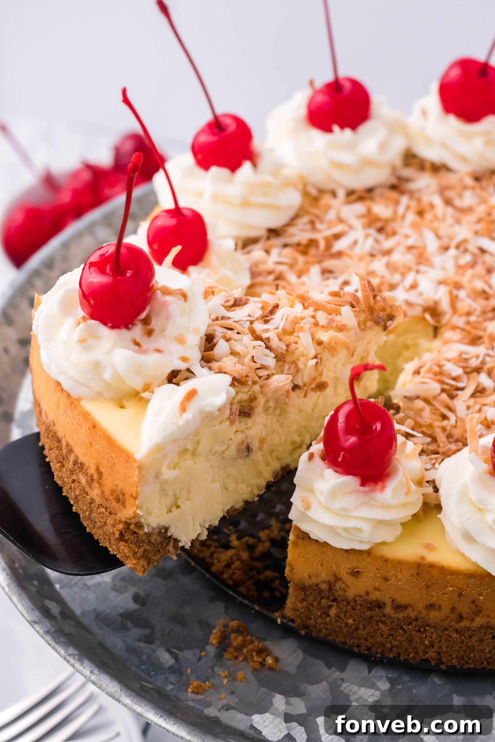 A black spatula lifting a pristine slice of Coconut Cheesecake from its silver cake dish, showcasing its perfect form.