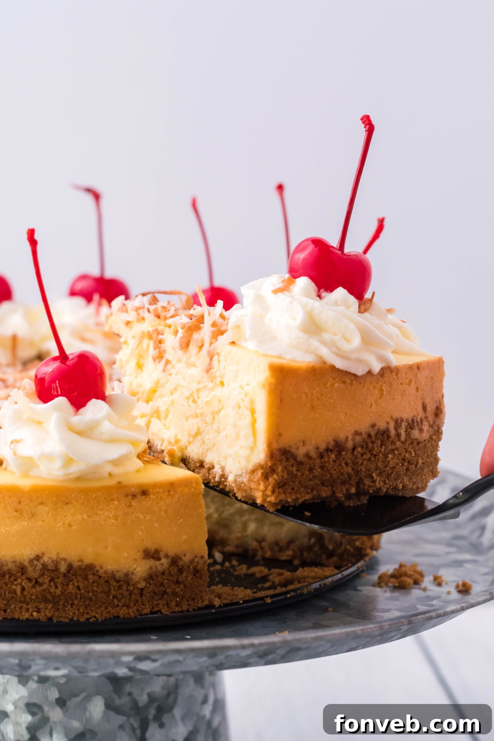 A slice of Coconut Cheesecake being carefully lifted from a silver cake dish with a black spatula, ready to be served.