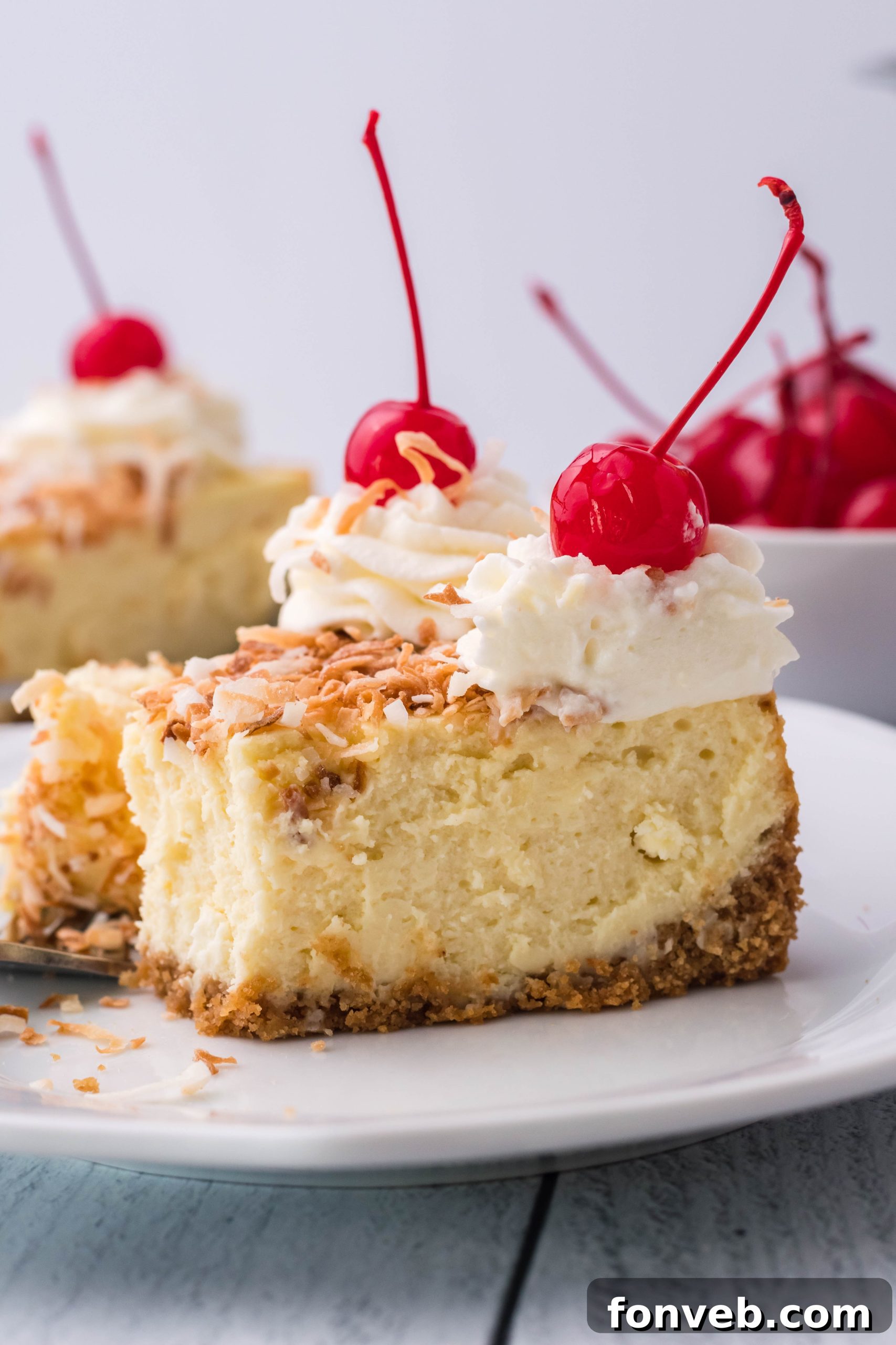 A close-up side shot of a Coconut Cheesecake on a white plate, beautifully garnished with bright red cherries and a dollop of whipped cream.