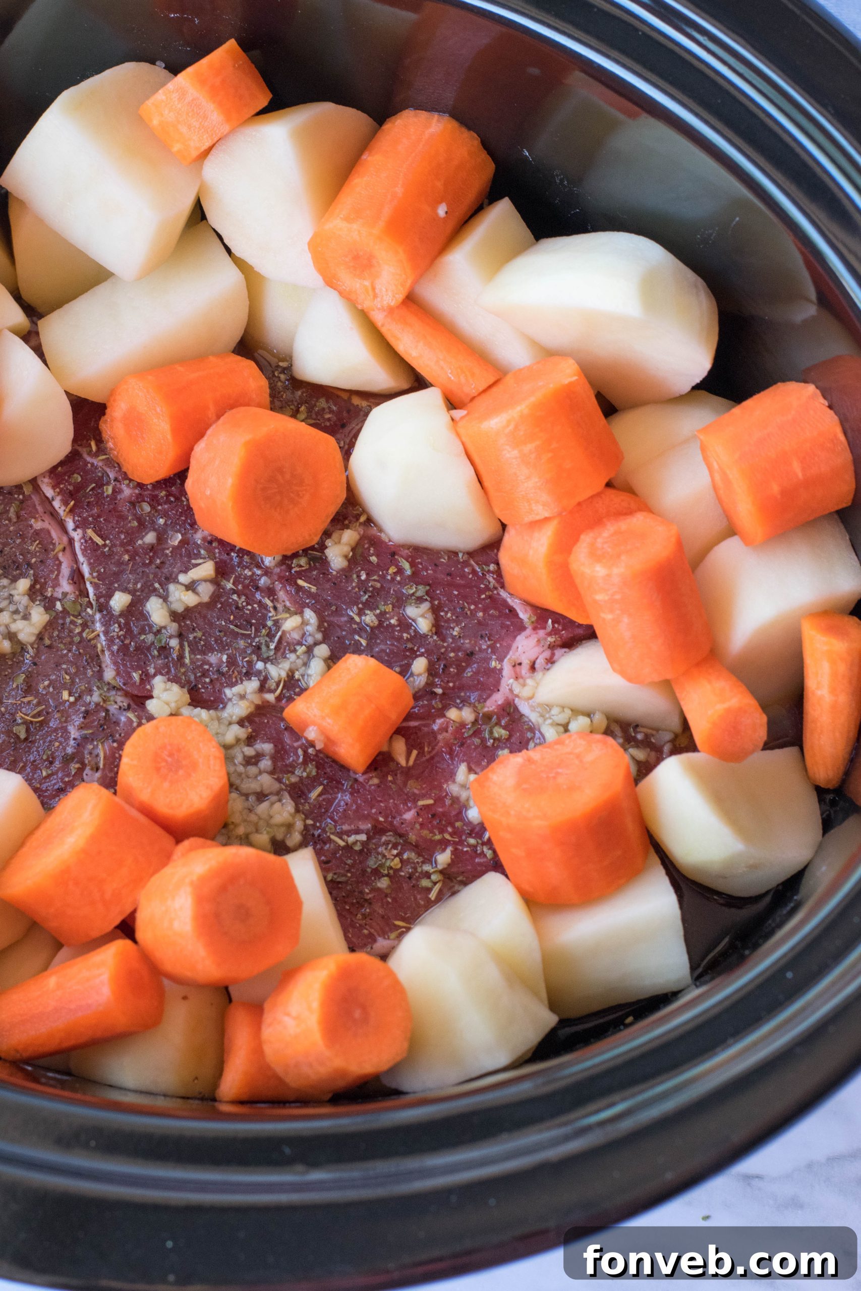 crock pot chuck roast in a black crock pot uncooked with chopped potatoes and carrots