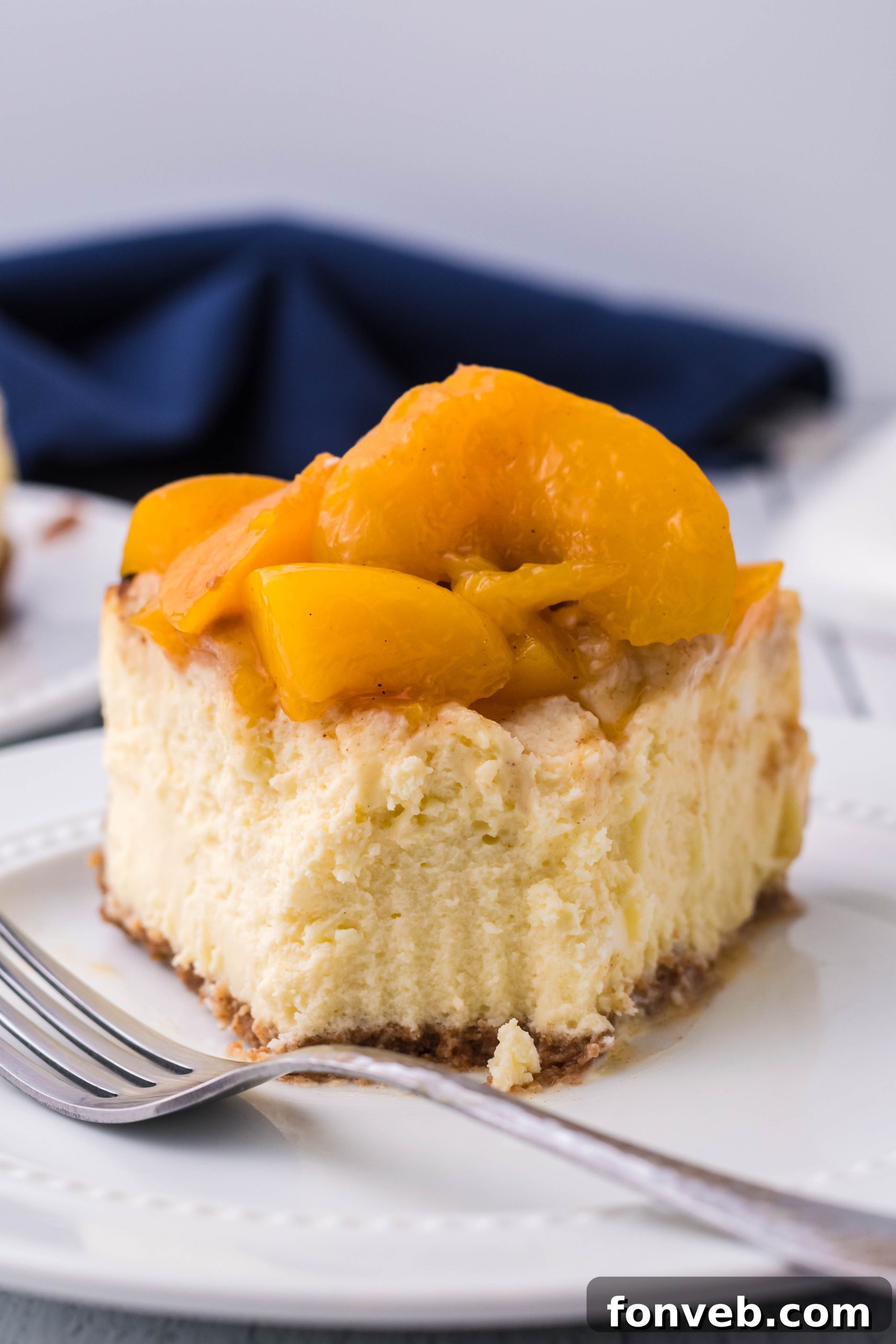 A delectable slice of Peach Cobbler Cheesecake on a white plate with a bite taken, revealing creamy texture and peach topping.