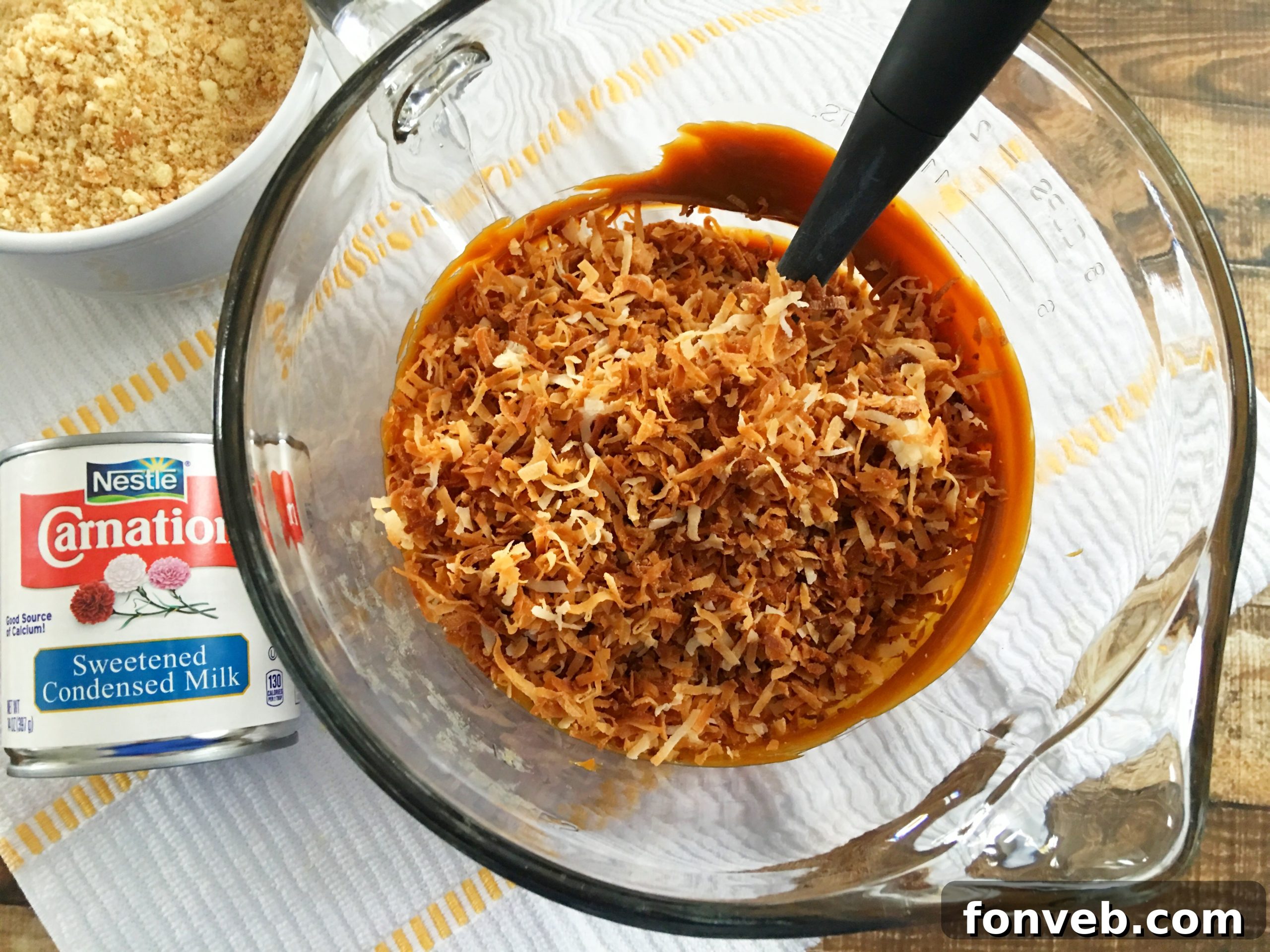Sweetened condensed milk being poured into a large glass mixing bowl, combining with the toasted coconut, dulce de leche, and crushed Nilla wafers for the Samoa Bite base.