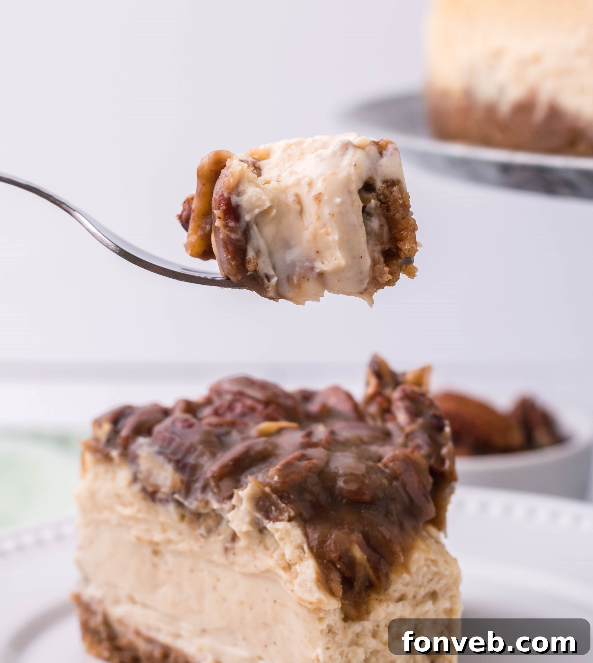 A slice of Pecan Pie Cheesecake on a white plate, with a bite taken and a fork lifting a piece, emphasizing its perfect texture.