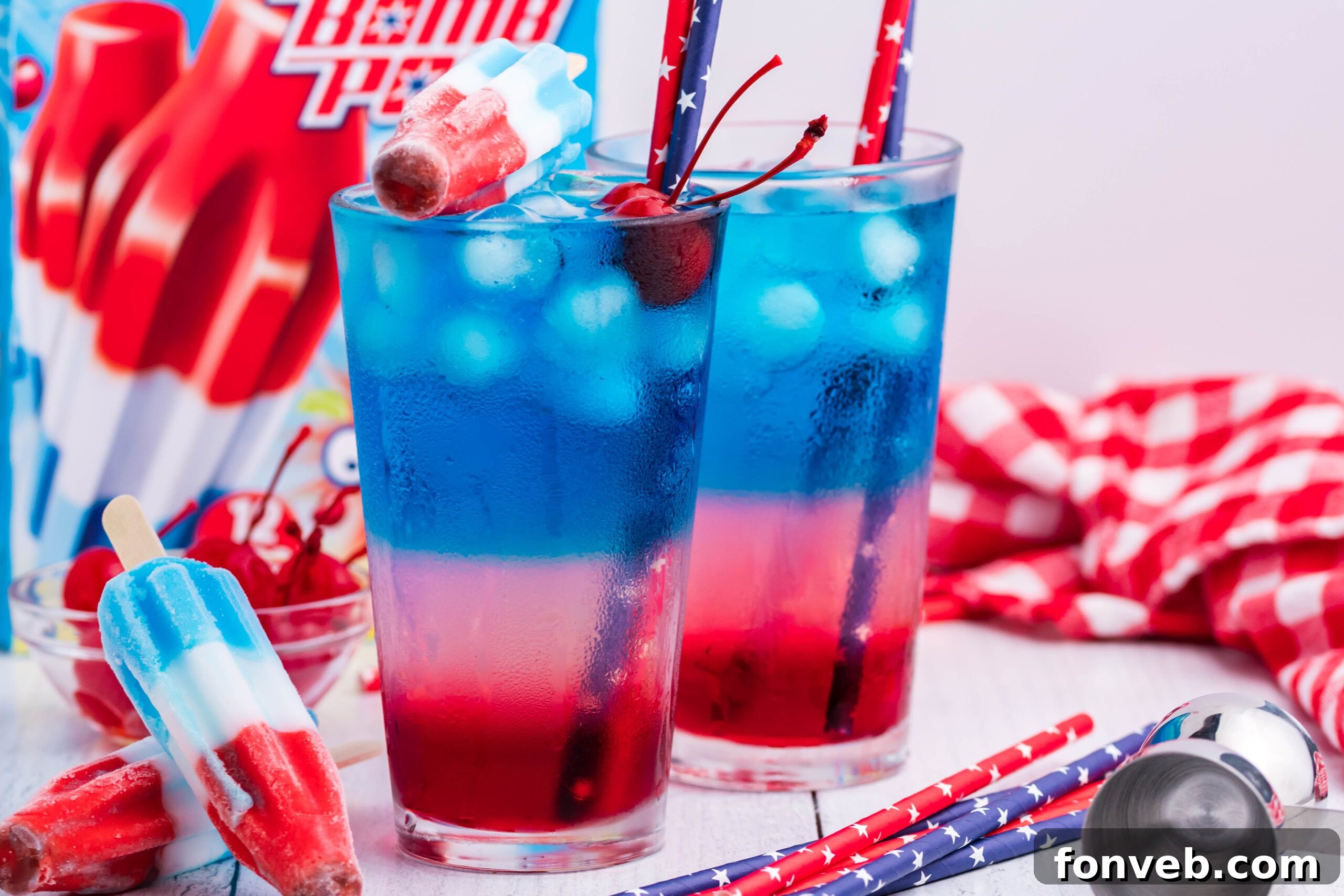 two Bomb Pop Cocktail Drink in a clear glass with a blue & red straw with cherries floating