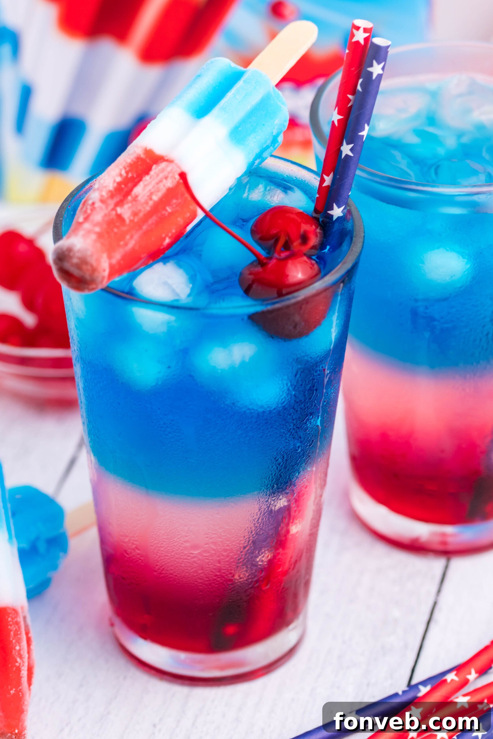 Bomb Pop Cocktail Drink in a clear glass with a blue & red straw with cherries floating and a ice cream sitting on top