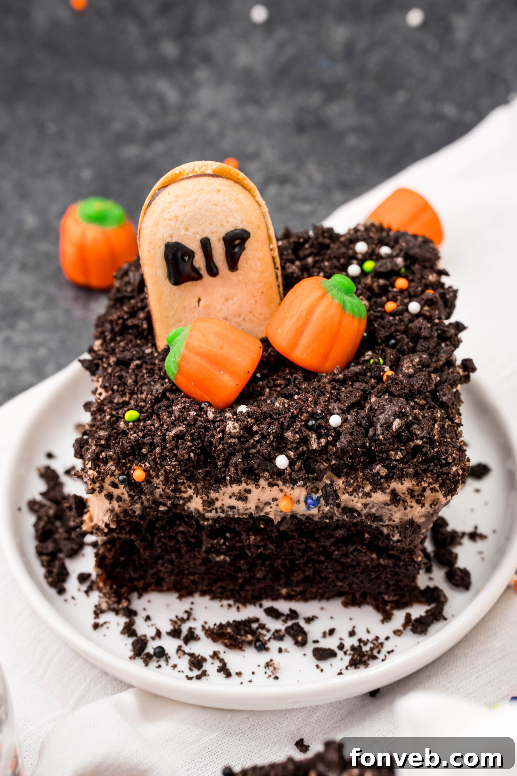 A delectable slice of Halloween Dirt Cake, richly adorned with Oreo crumbles and miniature candy pumpkins, presented on a clean white plate, offering a taste of spooky indulgence.