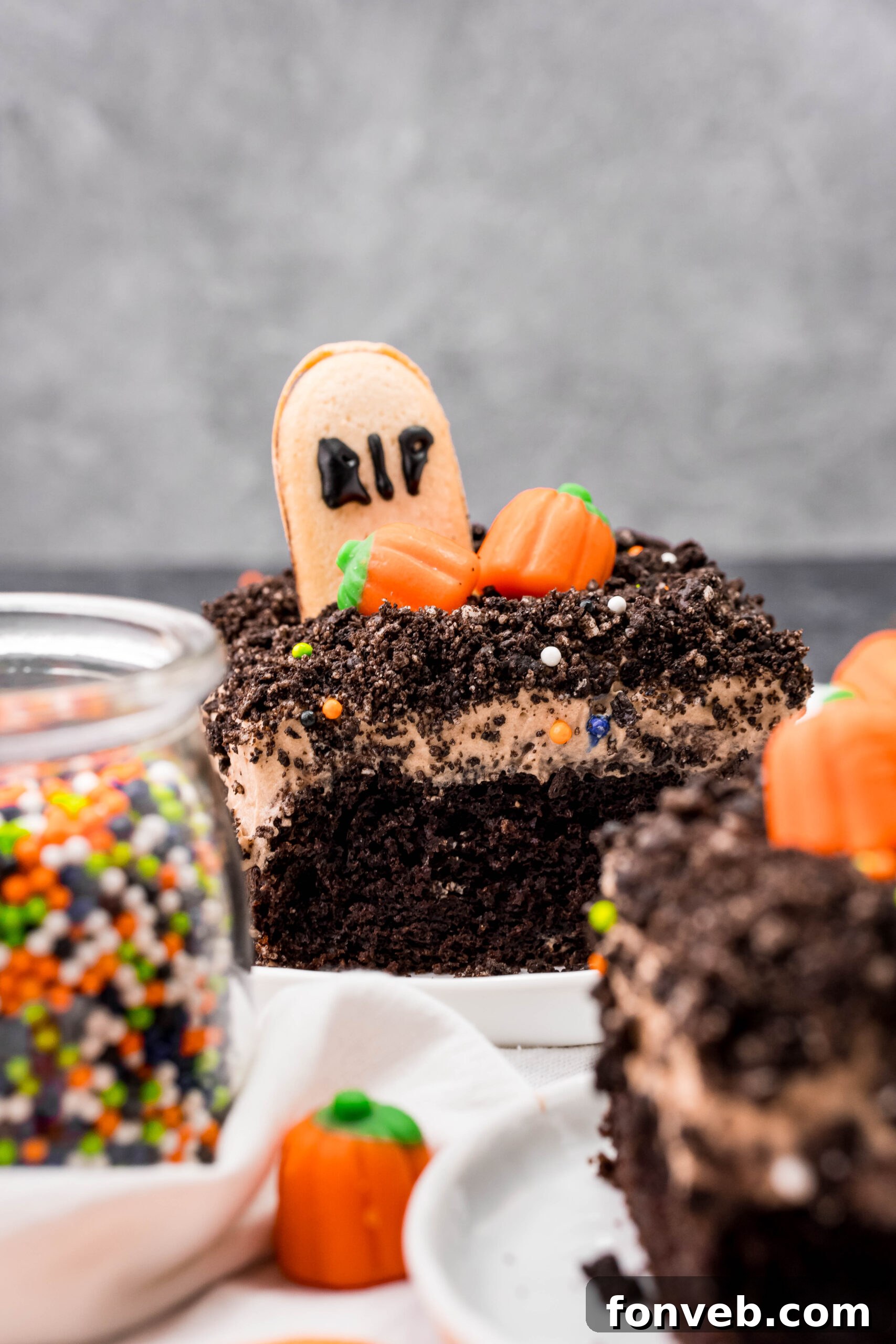 A delightful slice of Halloween Dirt Cake, presented on a white plate, with a glass jar filled with festive Halloween-colored sprinkles positioned invitingly beside it.