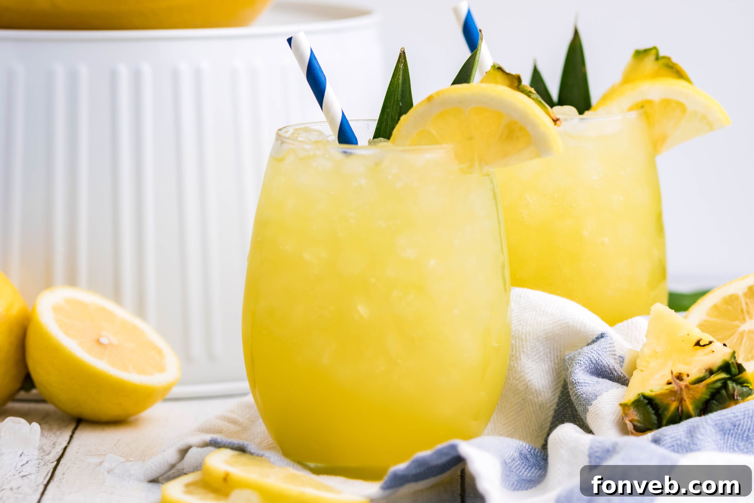 Pineapple Lemonade in a clear glass with ice, a blue striped straw, and a fresh lemon slice
