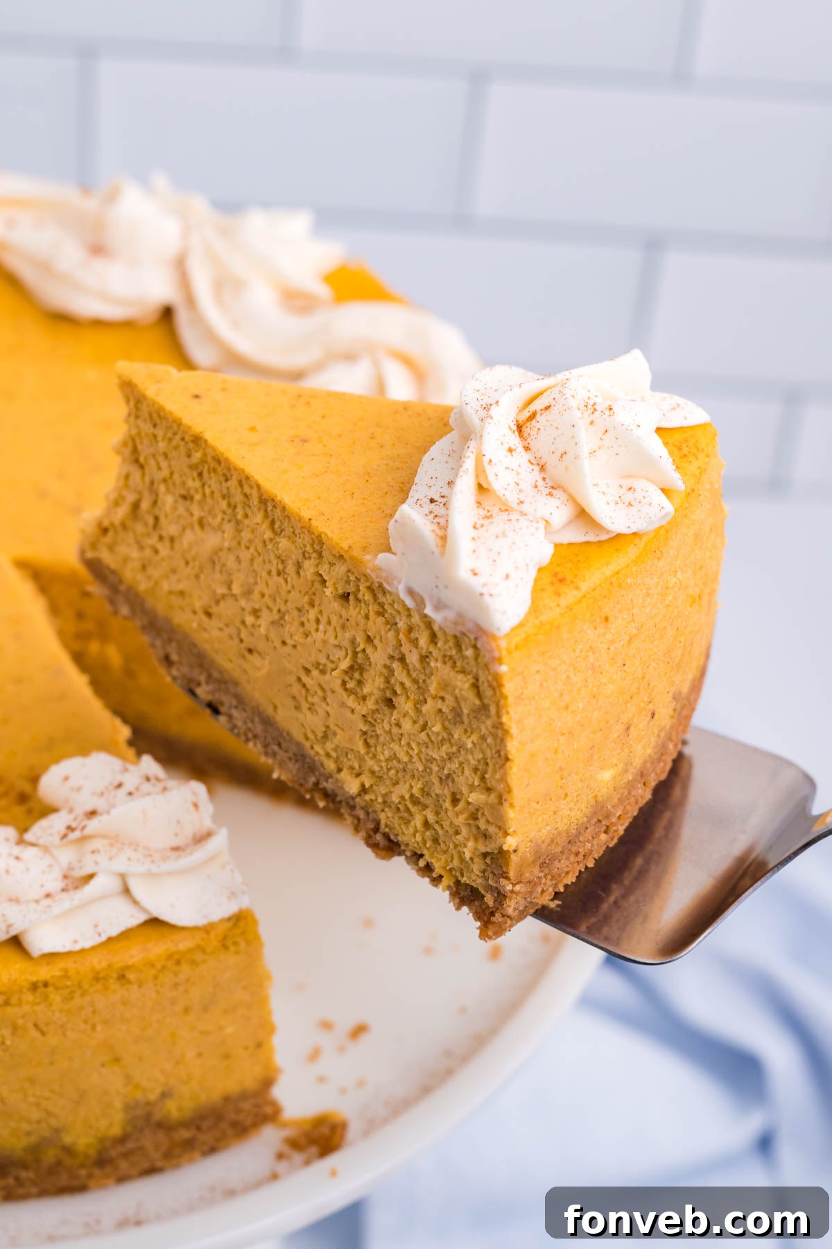 A slice of Pumpkin Cheesecake being lifted from the full cake on a white cake stand, showing its creamy texture