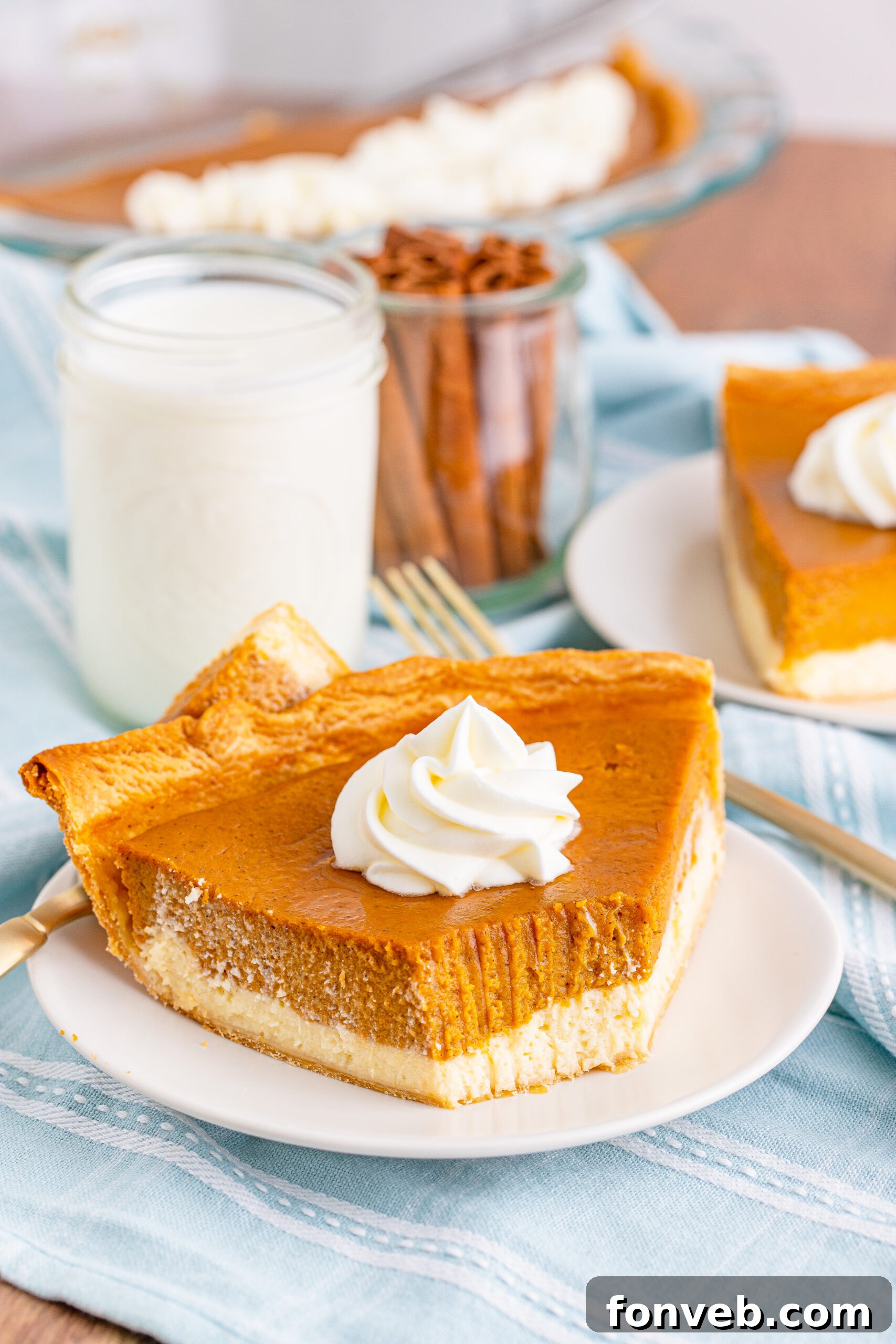 A slice of Pumpkin Pie Cheesecake on a white plate, accompanied by a glass of milk and cinnamon sticks in a jar in the background