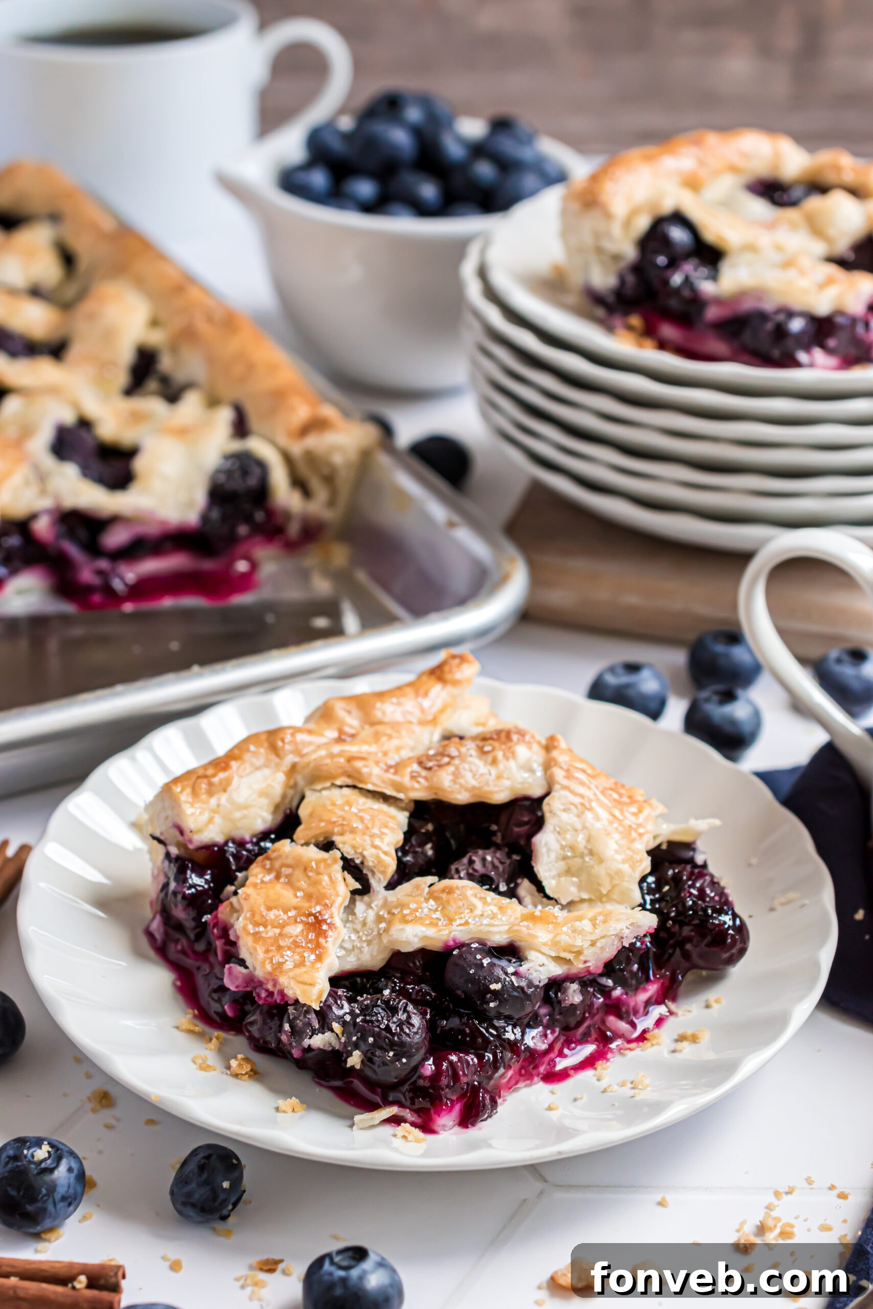 A slice of vibrant Blueberry Slab Pie on a white plate, garnished with fresh blueberries and cinnamon sticks in the background.