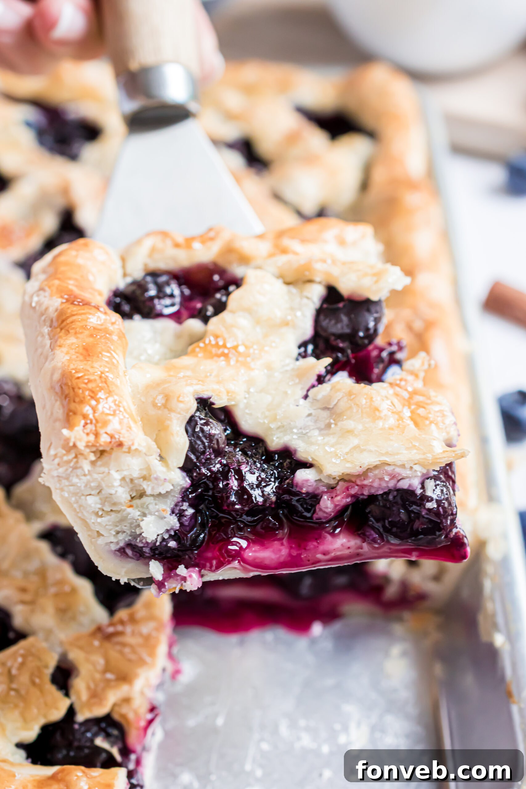 A golden-brown slice of blueberry pie being carefully removed from the baking pan using a silver and wooden spatula.