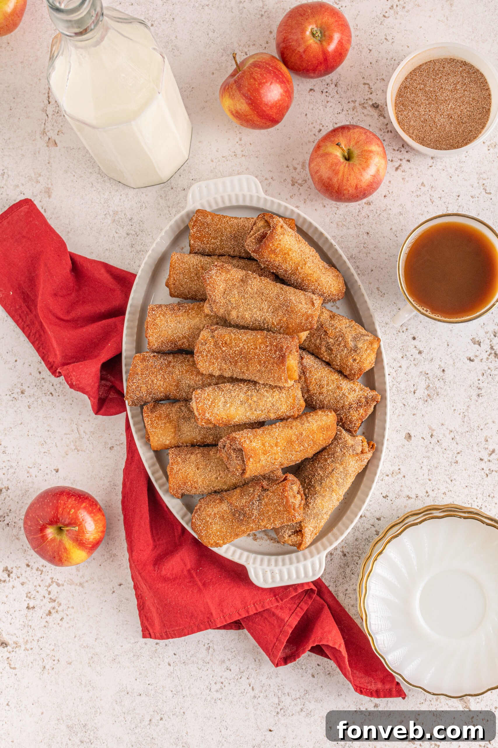 Overhead view of egg rolls stacked on a white platter with apples and a red linen in the background. 