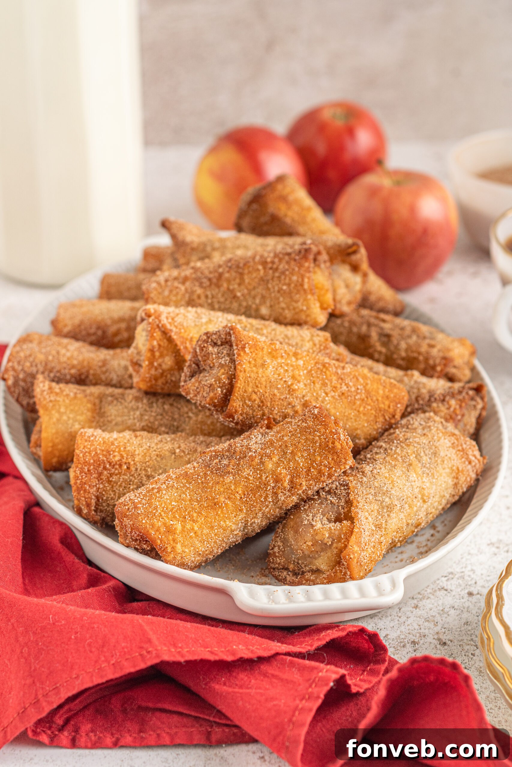 Apple pie egg rolls stacked on a white platter with apples and a red linen in the background. 