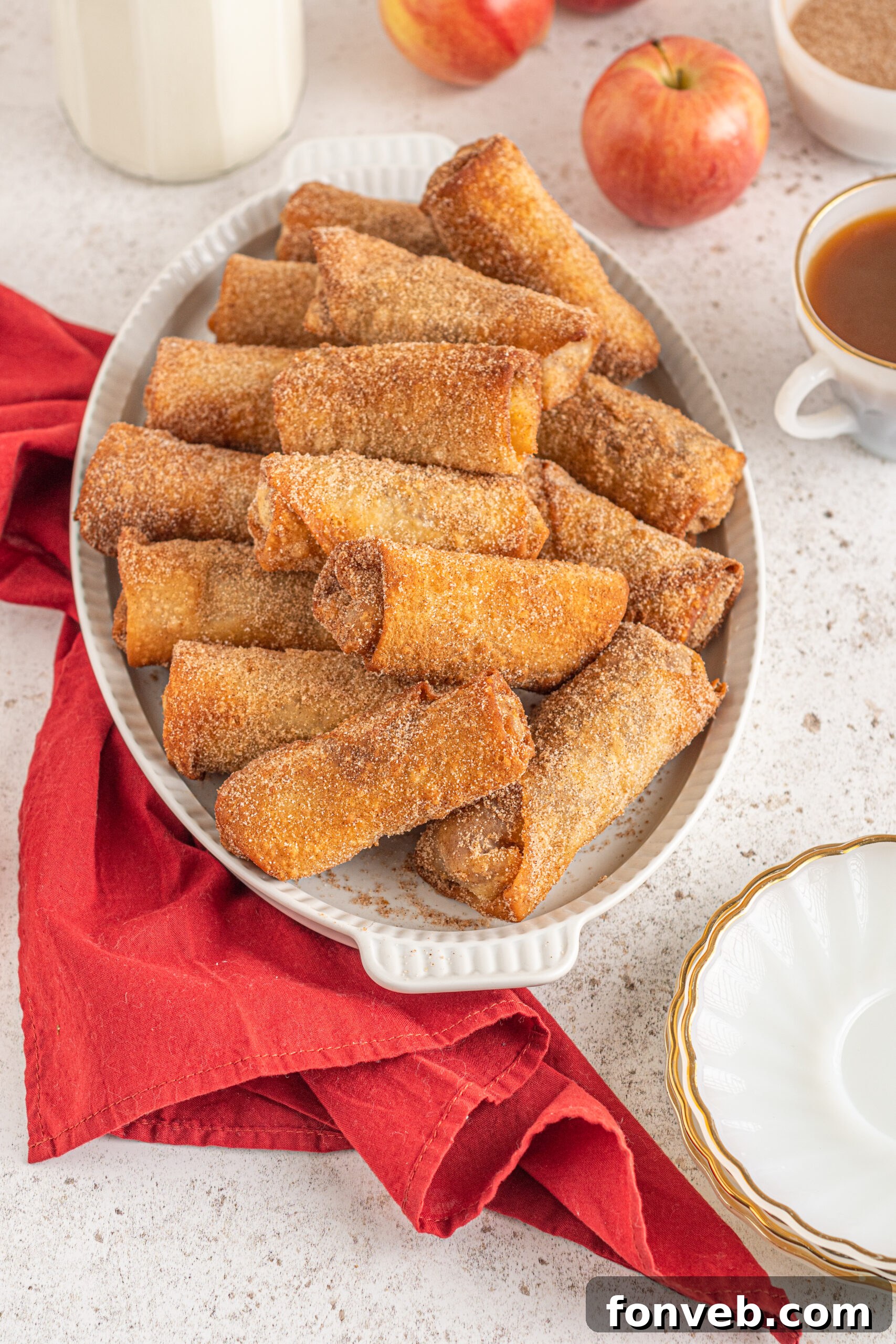 Egg rolls stacked on a white platter with apples and a red linen in the background. 
