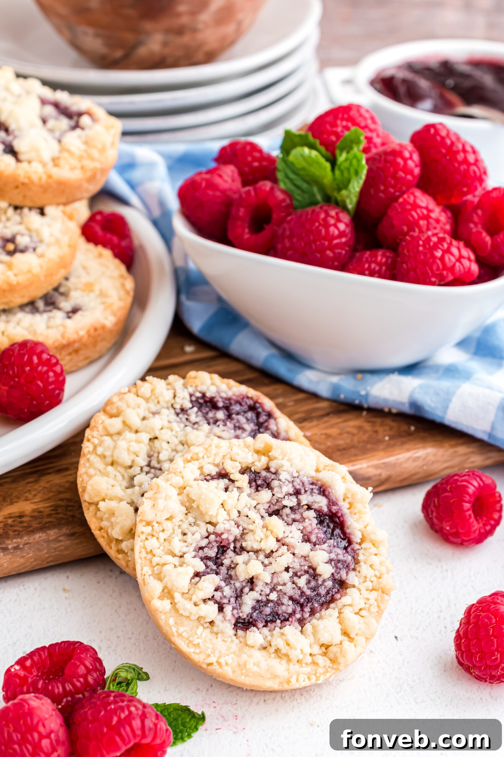 Two golden-baked raspberry cookies with a crumbly topping, propped against a rustic wooden cutting board, with fresh, vibrant raspberries scattered nearby in a white ceramic bowl.