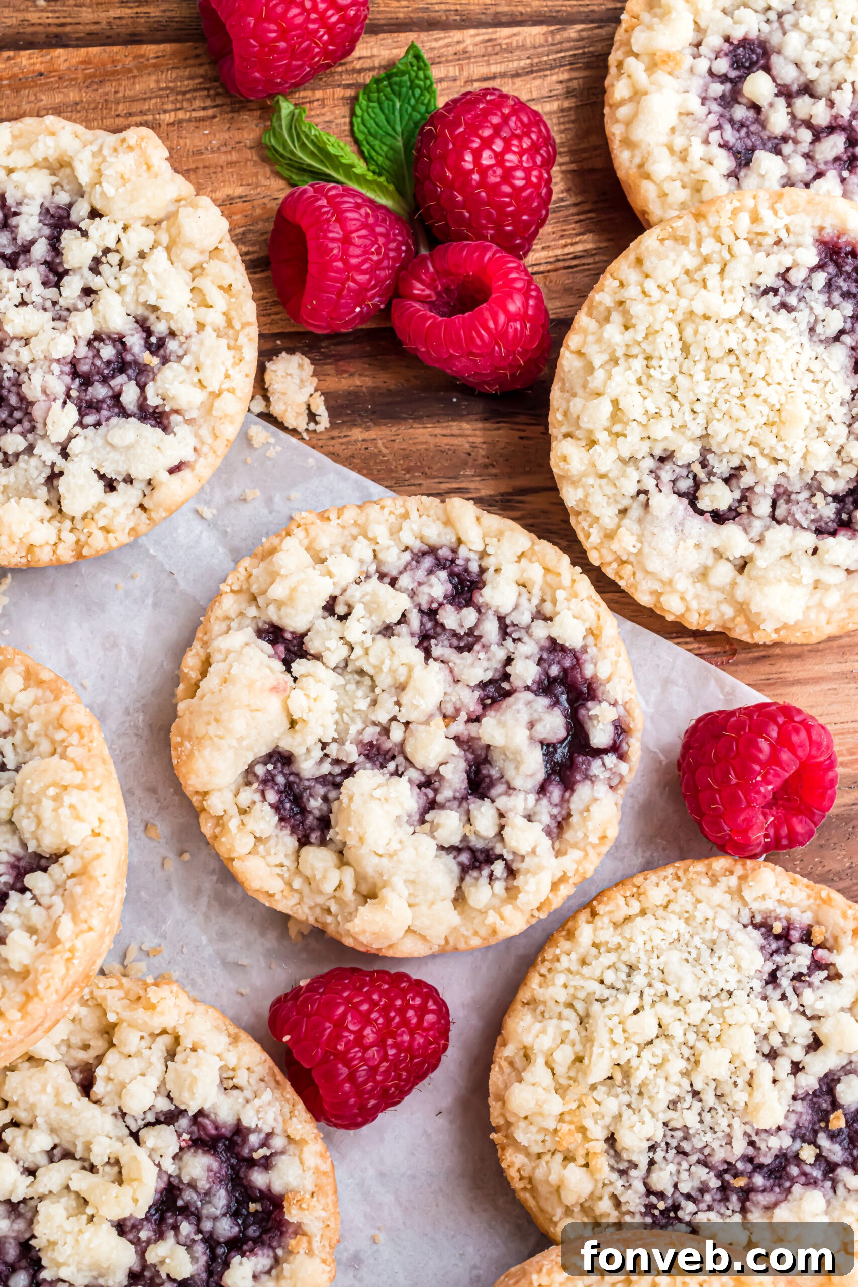 An overhead shot of several baked raspberry cookies arranged on wax paper, with fresh, vibrant raspberries scattered around them, suggesting readiness for storage or serving.