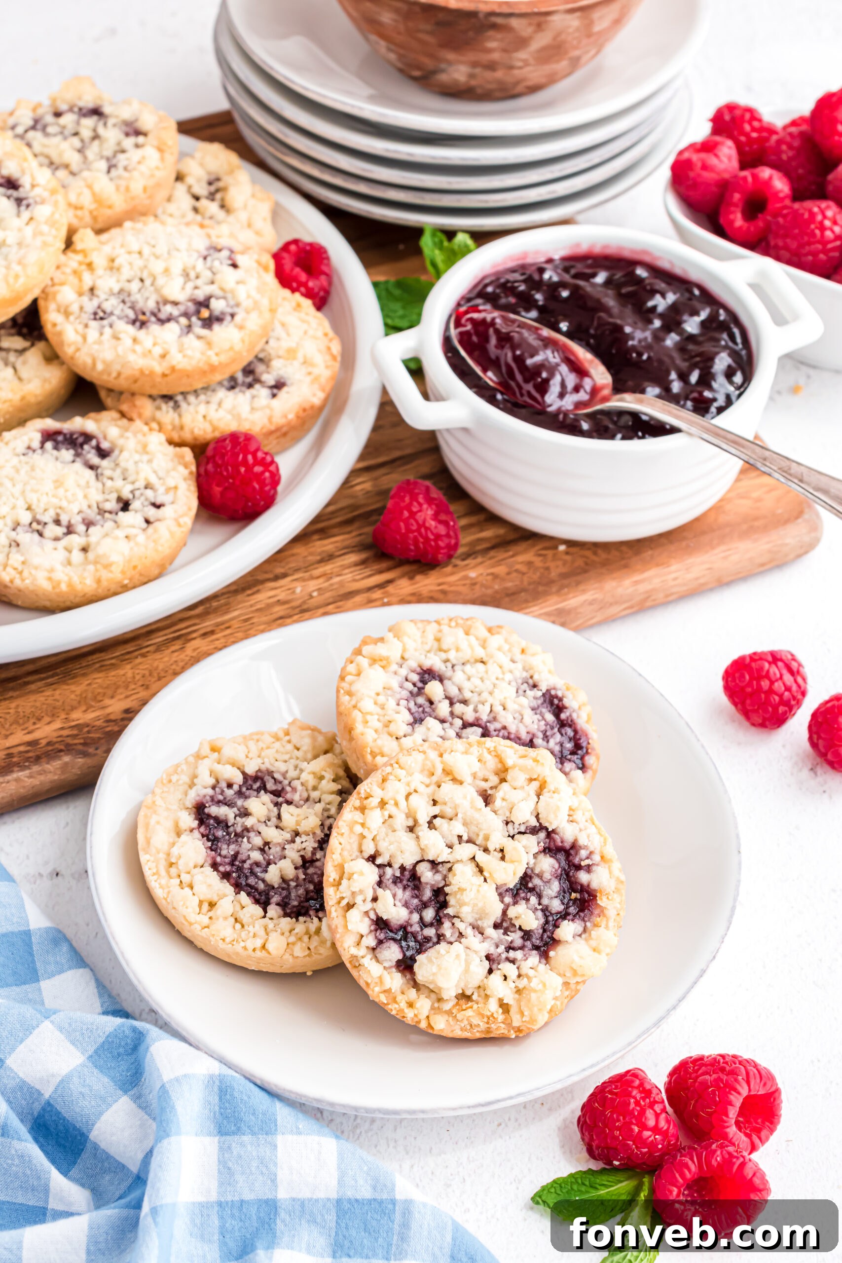 Three beautifully presented raspberry cookies on a white plate, accompanied by a blue and white patterned linen napkin and fresh raspberries in the soft background, creating an inviting scene.