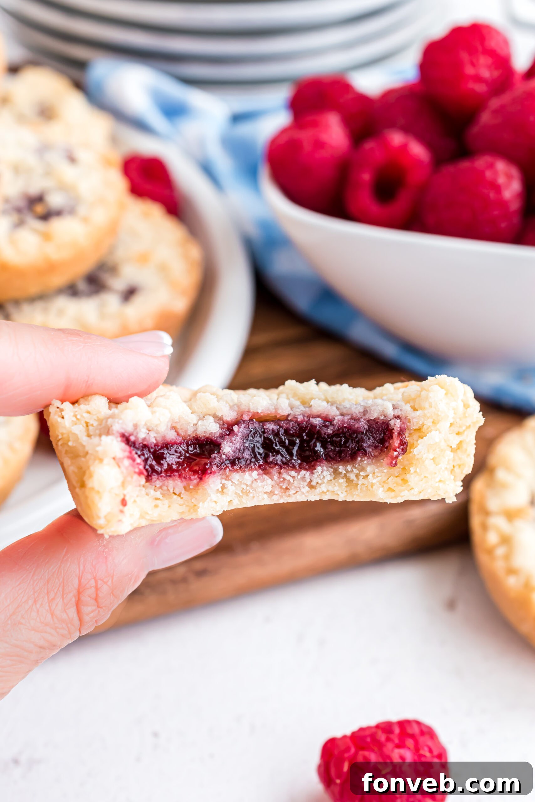 A close-up view of a raspberry cookie, cut in half to reveal the vibrant red raspberry jam filling and the delicate crumbly texture of the cookie and topping.