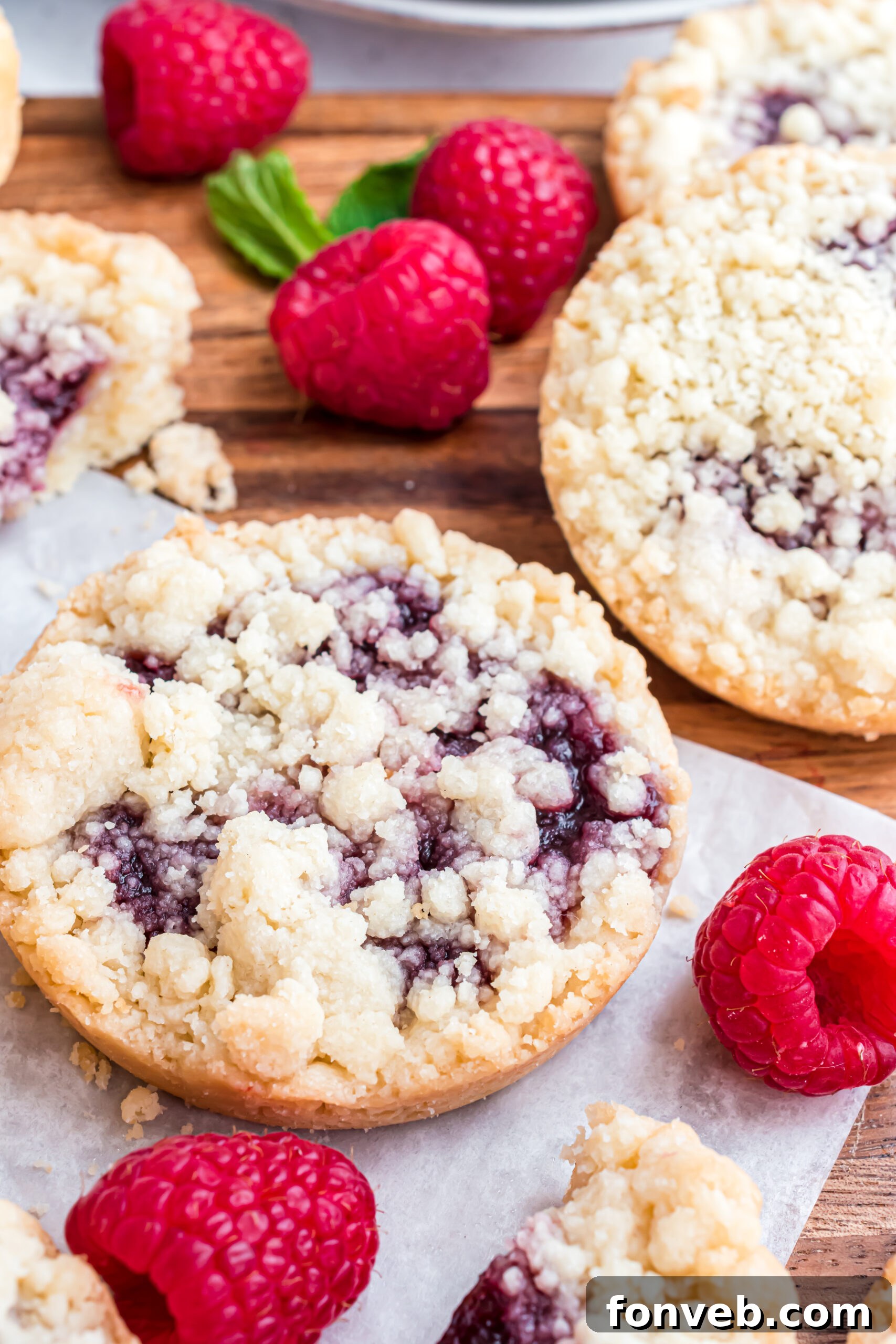 Several freshly baked raspberry cookies arranged neatly on a smooth wooden cutting board, showcasing their golden-brown crumble tops and hints of red jam.