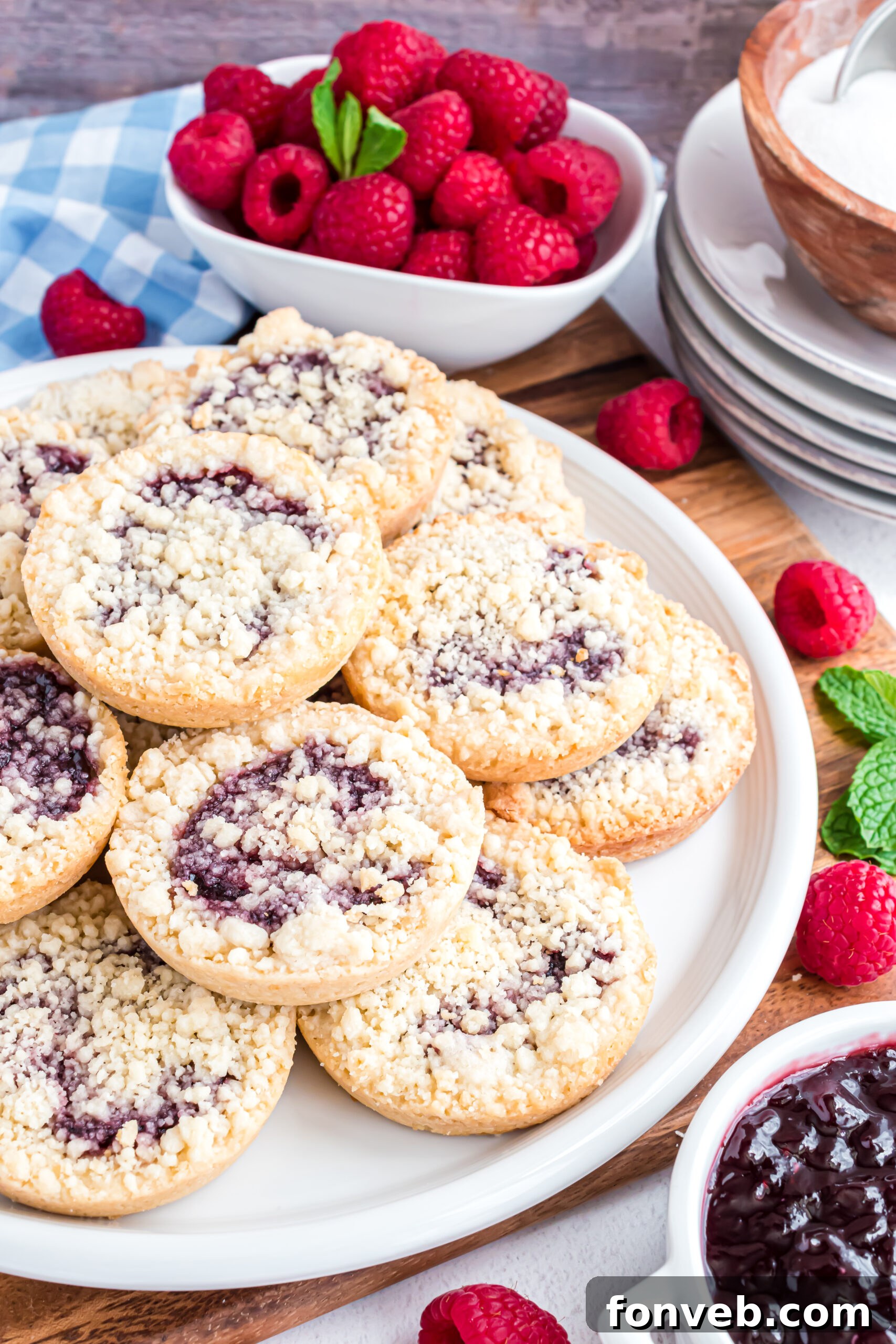 A stack of golden raspberry cookies on a pristine white plate, set on a wooden cutting board, with a scattering of fresh, ripe raspberries in the soft-focused background, emphasizing the fruitiness of the dessert.