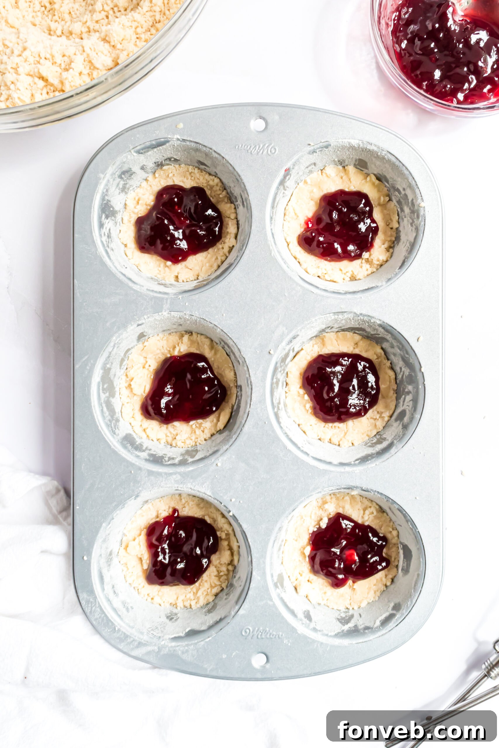 An overhead shot showcasing dollops of vibrant red raspberry jam carefully spooned into the center of each cookie crust in the muffin tin.