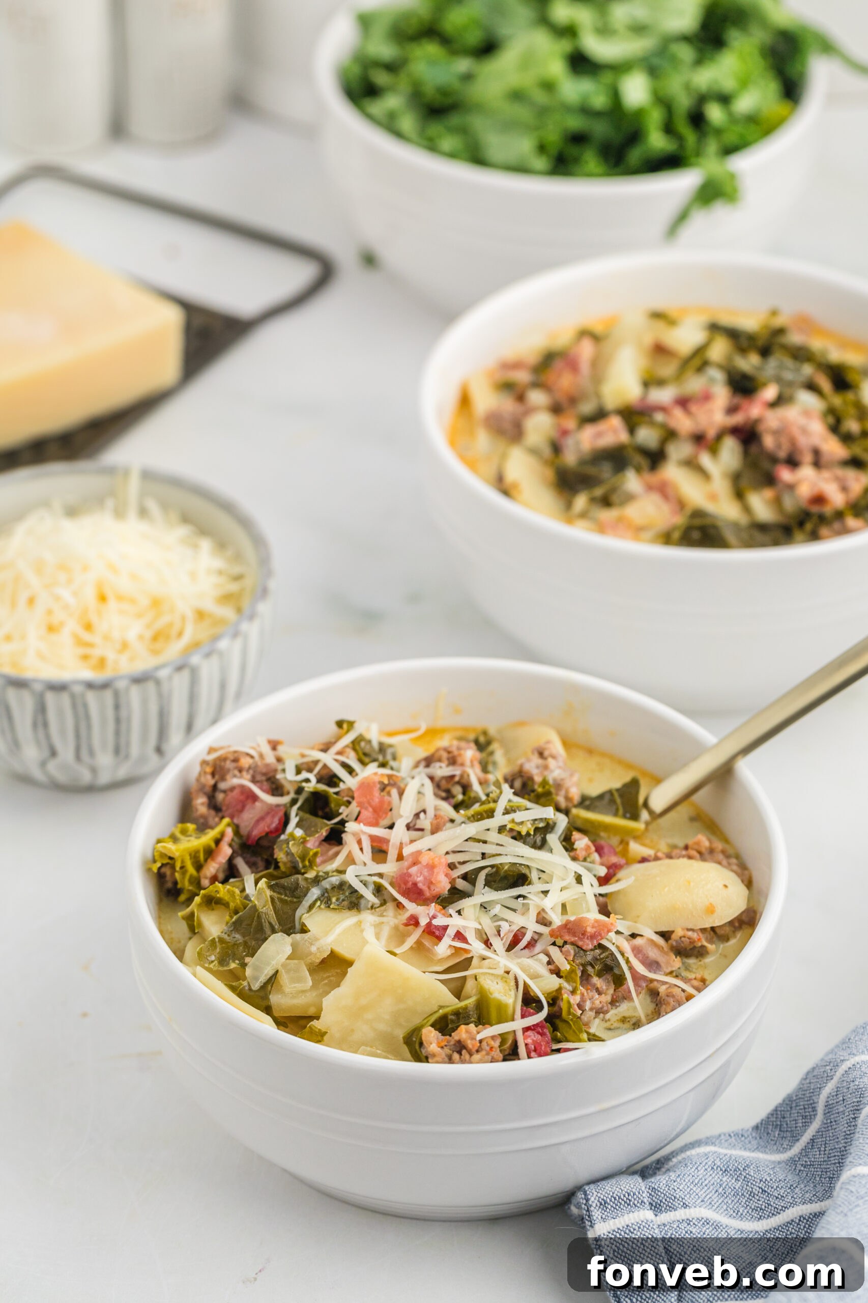 45 degree angle view of the Toscana soup in a white bowl topped with shredded cheese. Shredded cheese and kale in a white bowl in the background.  