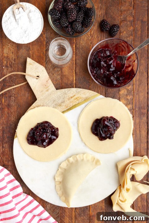 three circle of pie dough on a marble serving tray with blackberry jam in the center of each circle. One is folded in half showing the correct way to fold a hand pie.