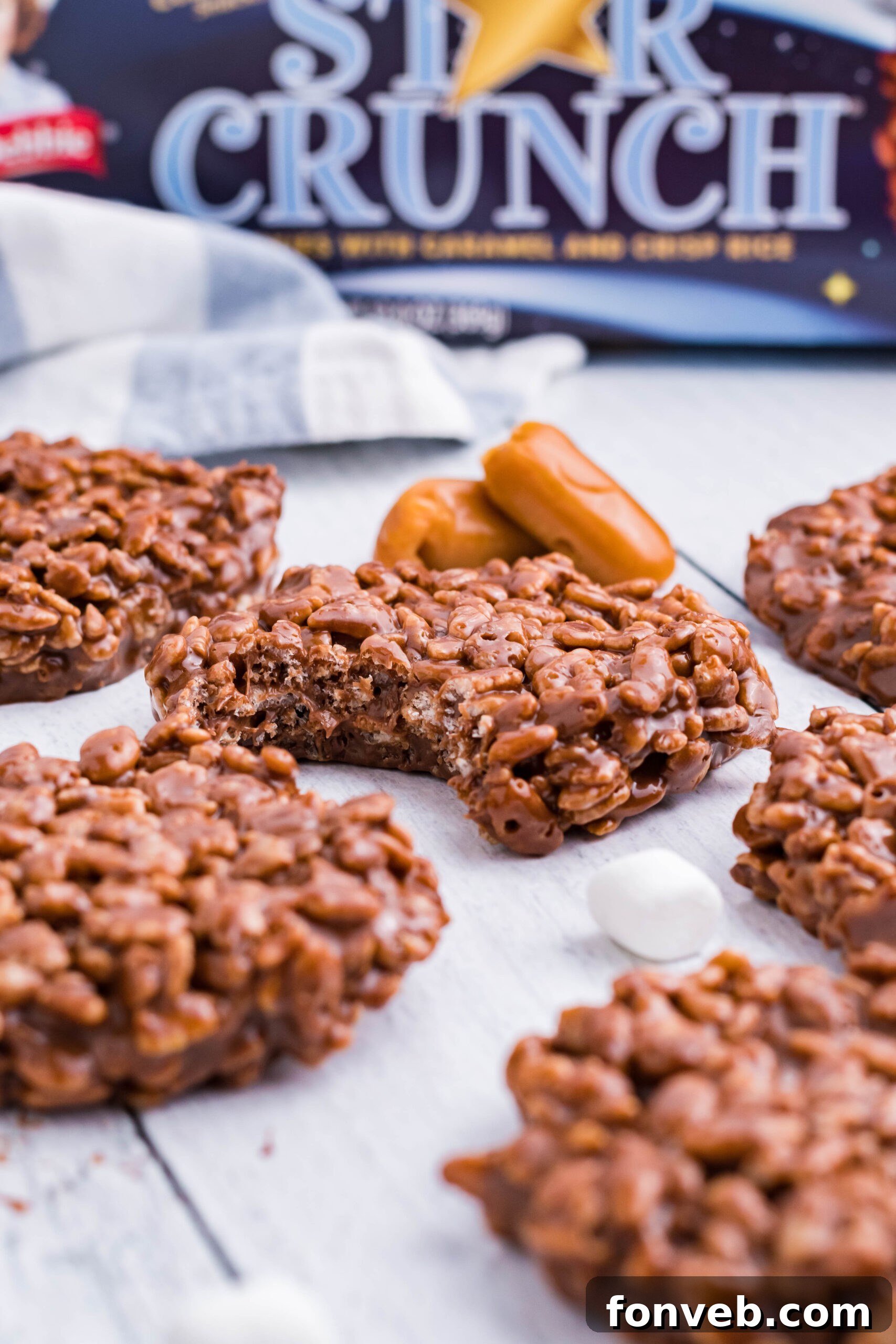 A single, perfectly formed Star Crunch copycat cookie sits on a rustic white wooden surface, with a small section bitten away, revealing the delectable layers of crispy rice cereal, soft caramel, and rich chocolate within.