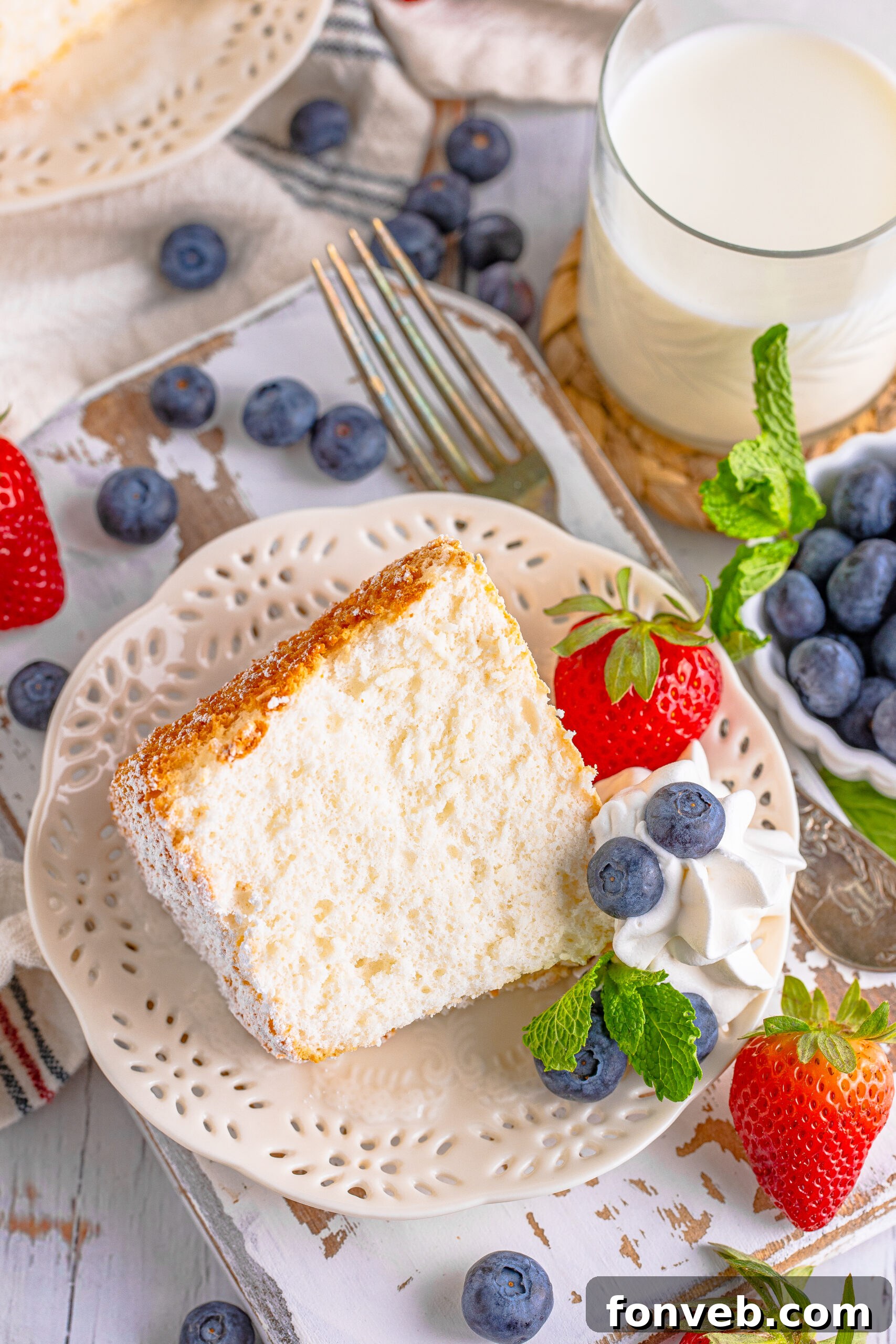 A slice of homemade Angel Food Cake on a white plate, elegantly garnished with whipped cream, fresh red berries, and a sprig of mint, highlighting its light texture.