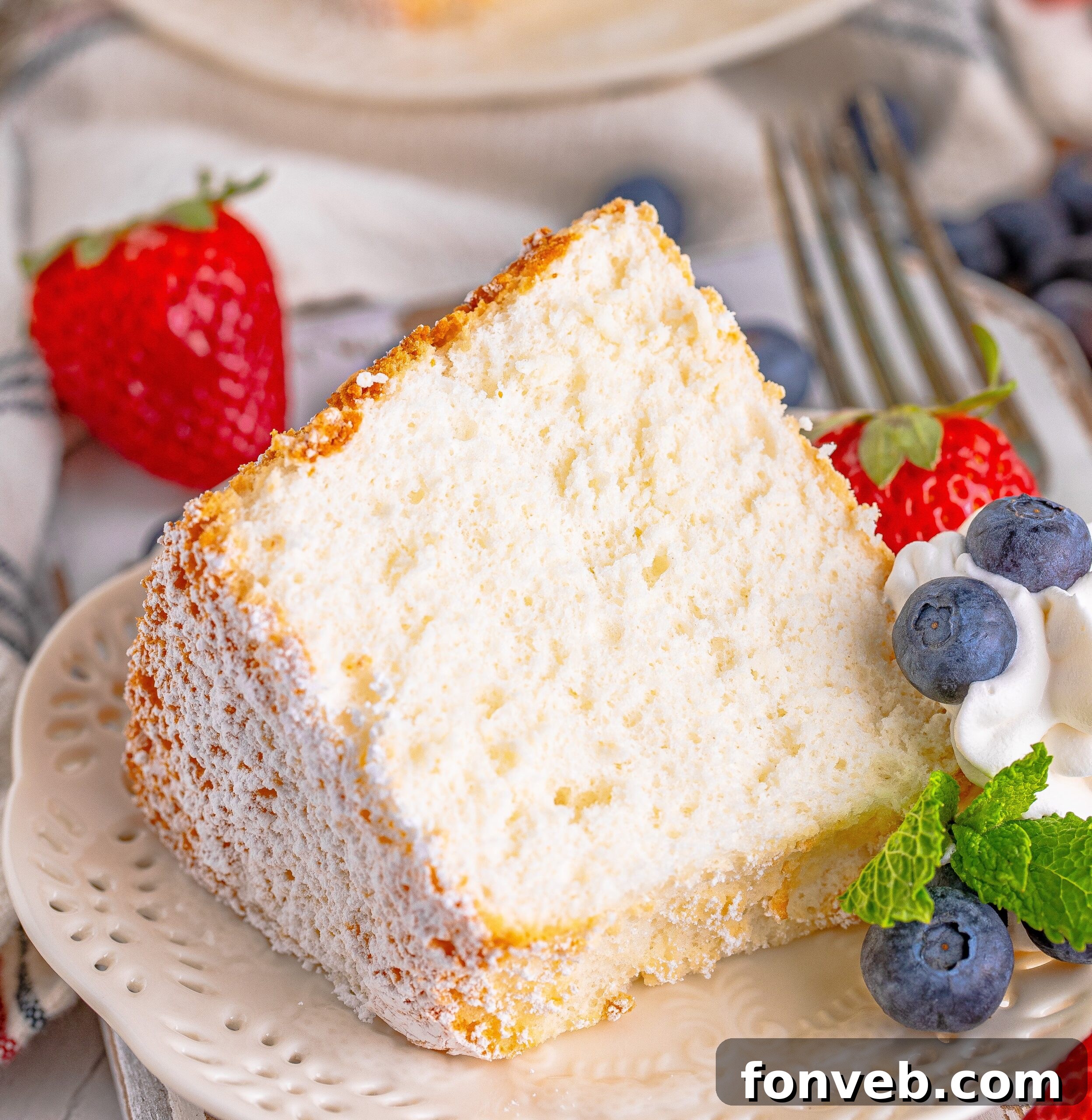 A close-up view of a slice of Angel Food Cake, beautifully presented on a white plate with fresh whipped cream, an assortment of berries, and a mint sprig.