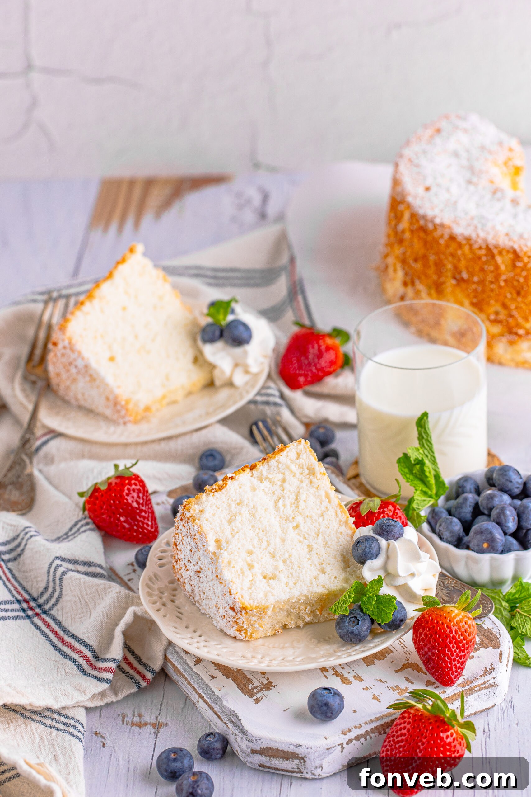 Two generous slices of Angel Food Cake served on a single white plate, each adorned with whipped cream, fresh mixed berries, and a mint garnish.